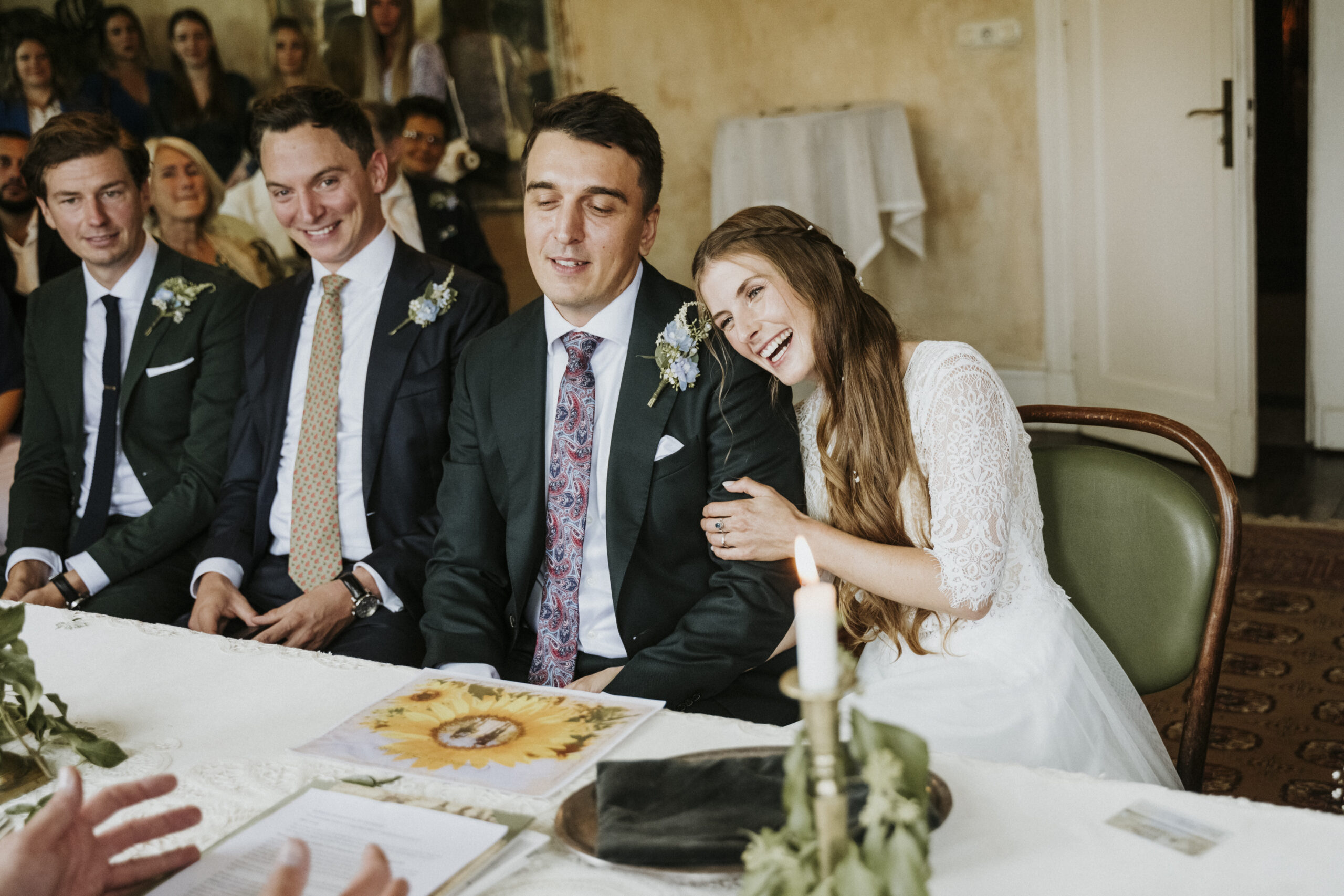 Hochzeitspaar mit Braut lächelnd am Tisch, Gäste im Hintergrund, Kerze und Dekoration vorne.