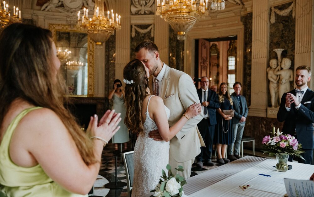 Hochzeitspaar küsst sich in elegantem Raum mit applaudierenden Gästen und Kronleuchtern.