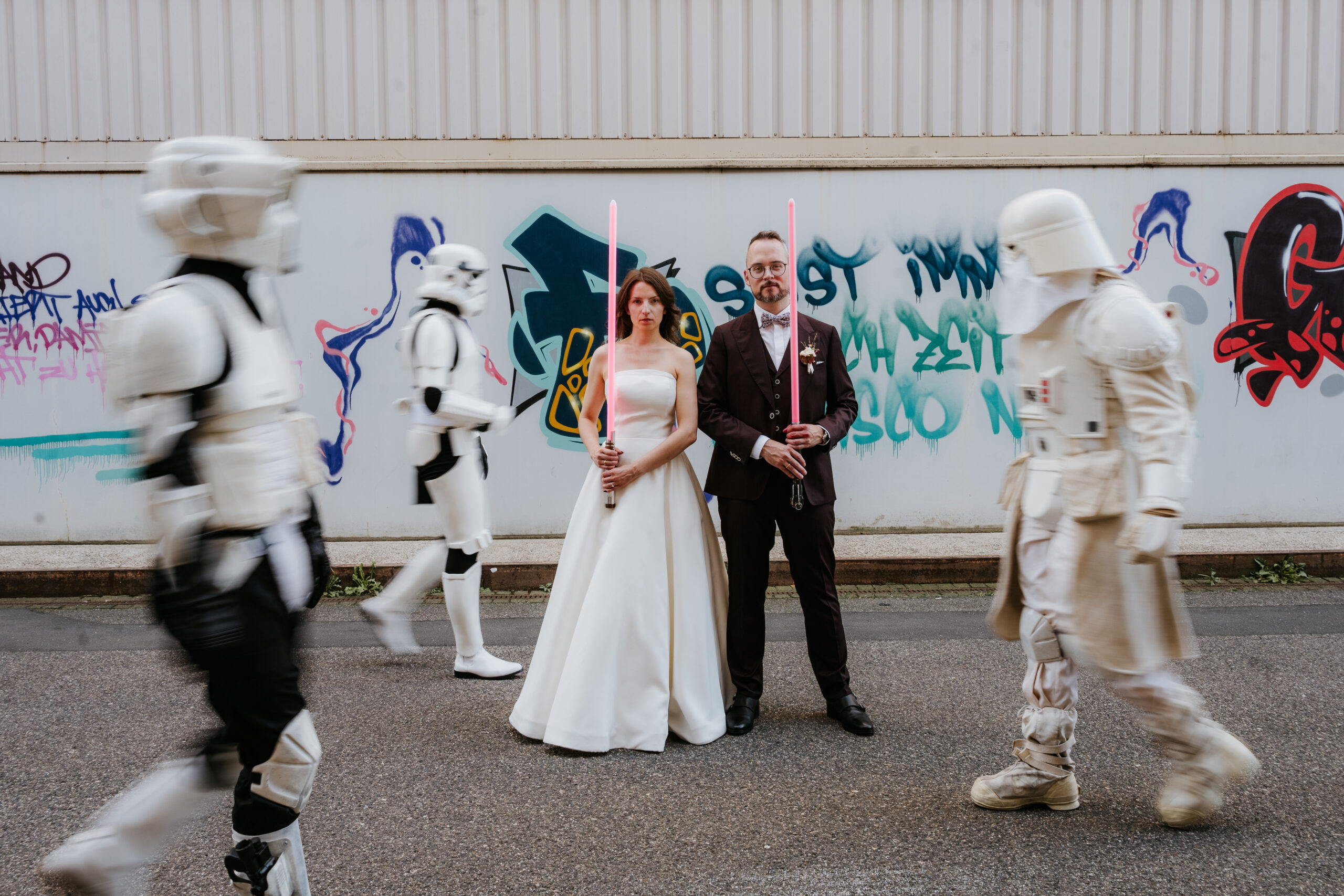 Hochzeitspaar mit Lichtschwertern, umgeben von Stormtroopern, vor Graffiti-Wand.