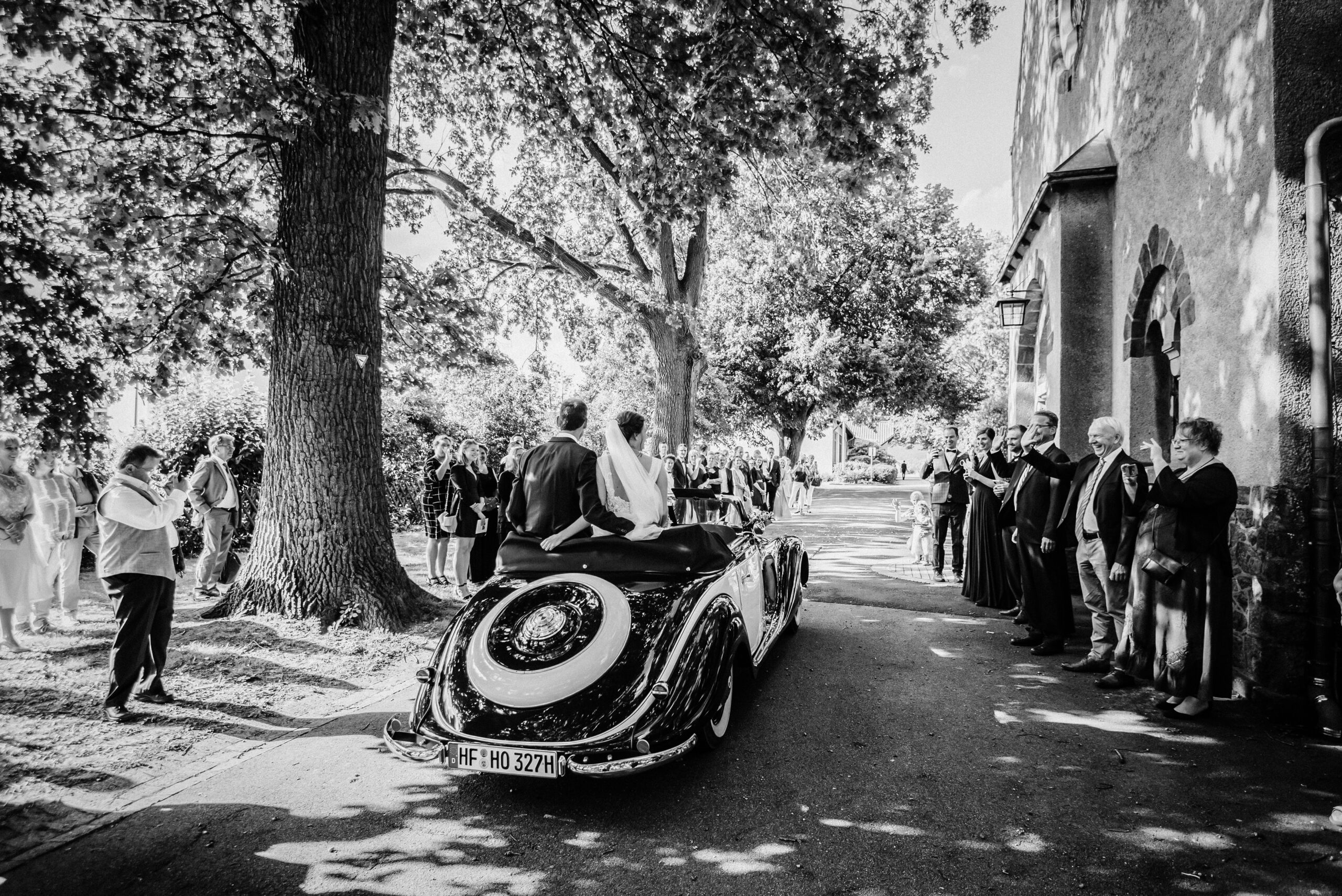 Hochzeitspaar im Oldtimer-Cabrio, von Gästen auf baumgesäumter Straße beobachtet.