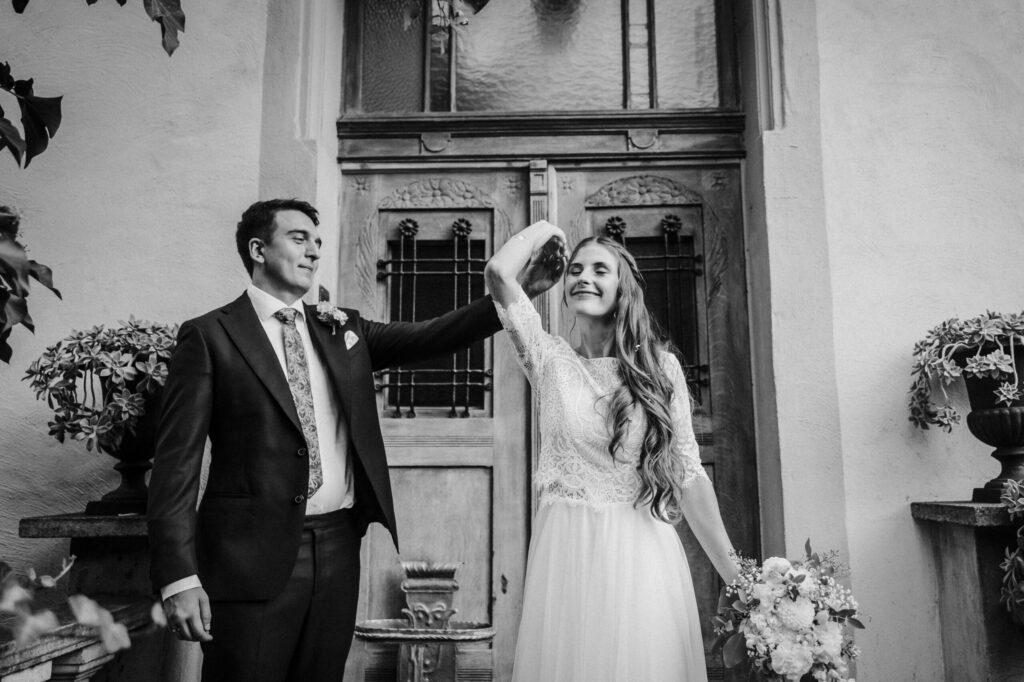 Hochzeitspaar tanzt vor alter Holztür, Mann im Anzug, Frau im Kleid mit Blumenstrauß.