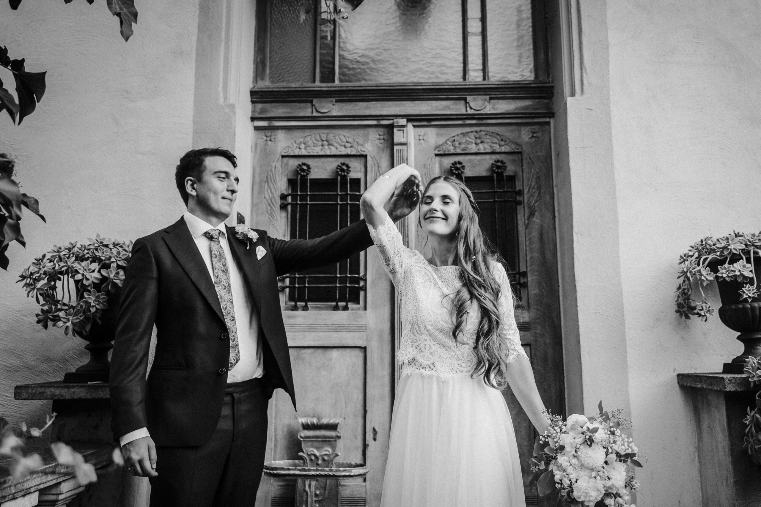 Hochzeitspaar tanzt vor alter Holztür, Mann im Anzug, Frau im Kleid mit Blumenstrauß.