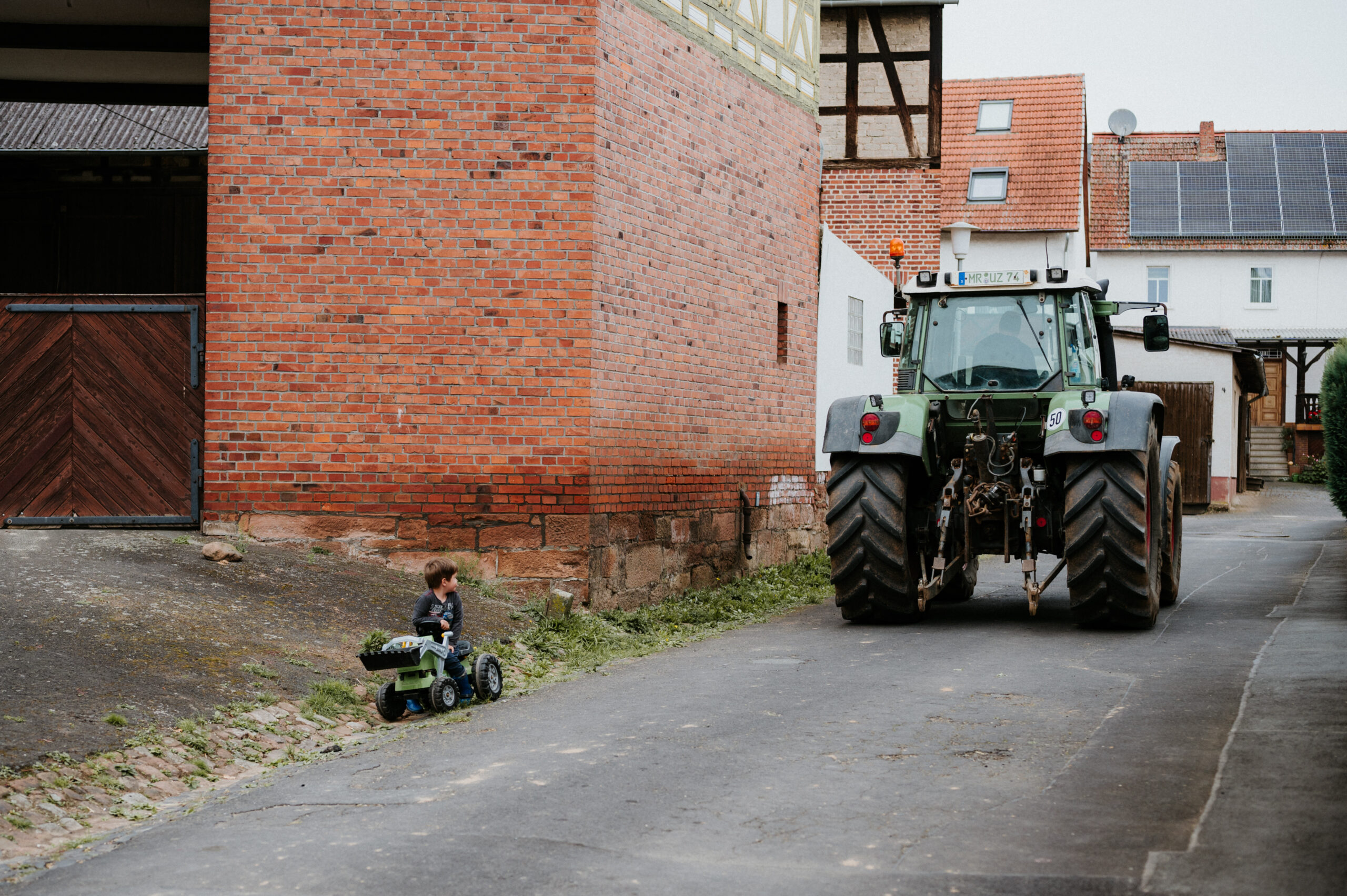 Kleiner Junge auf Spielzeugtraktor in Gasse, echter Traktor, rote Ziegel und Fachwerkhäuser.