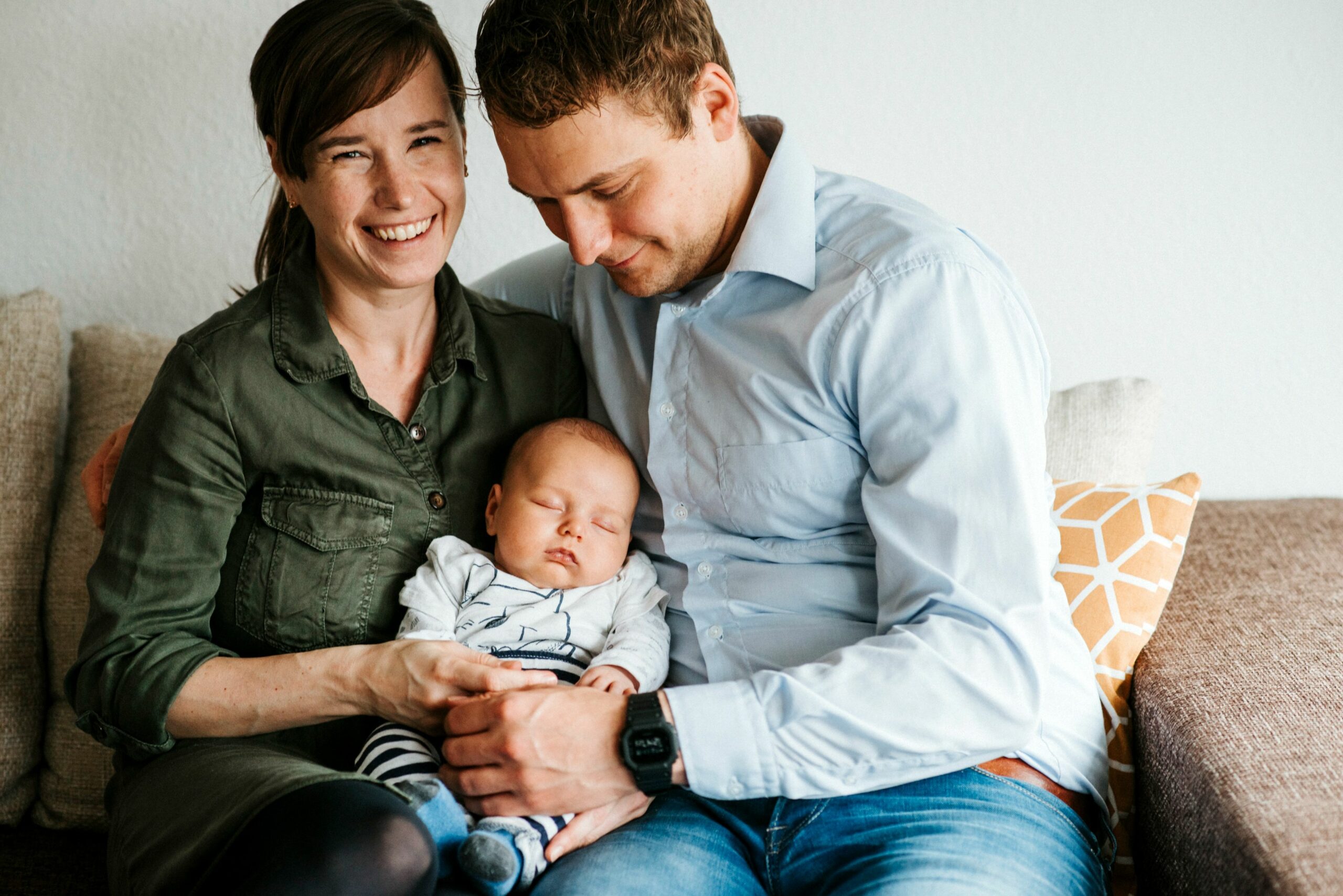 Lachende Frau und ein Mann auf Sofa halten schlafendes Baby in ihren Armen.