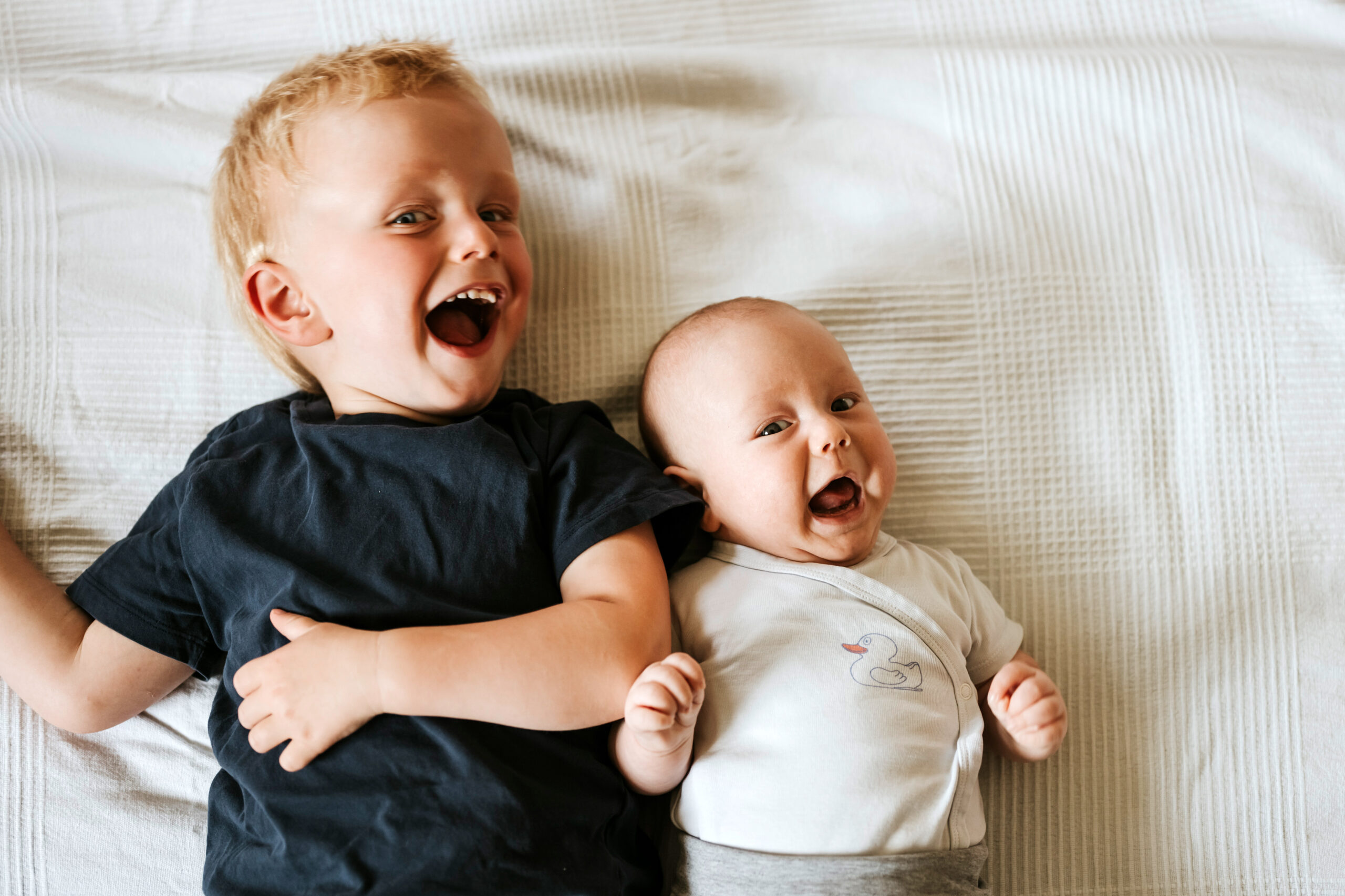 Zwei lachende Kinder auf einem weißen Bett, das ältere trägt ein dunkles T-Shirt, das Baby weiß.