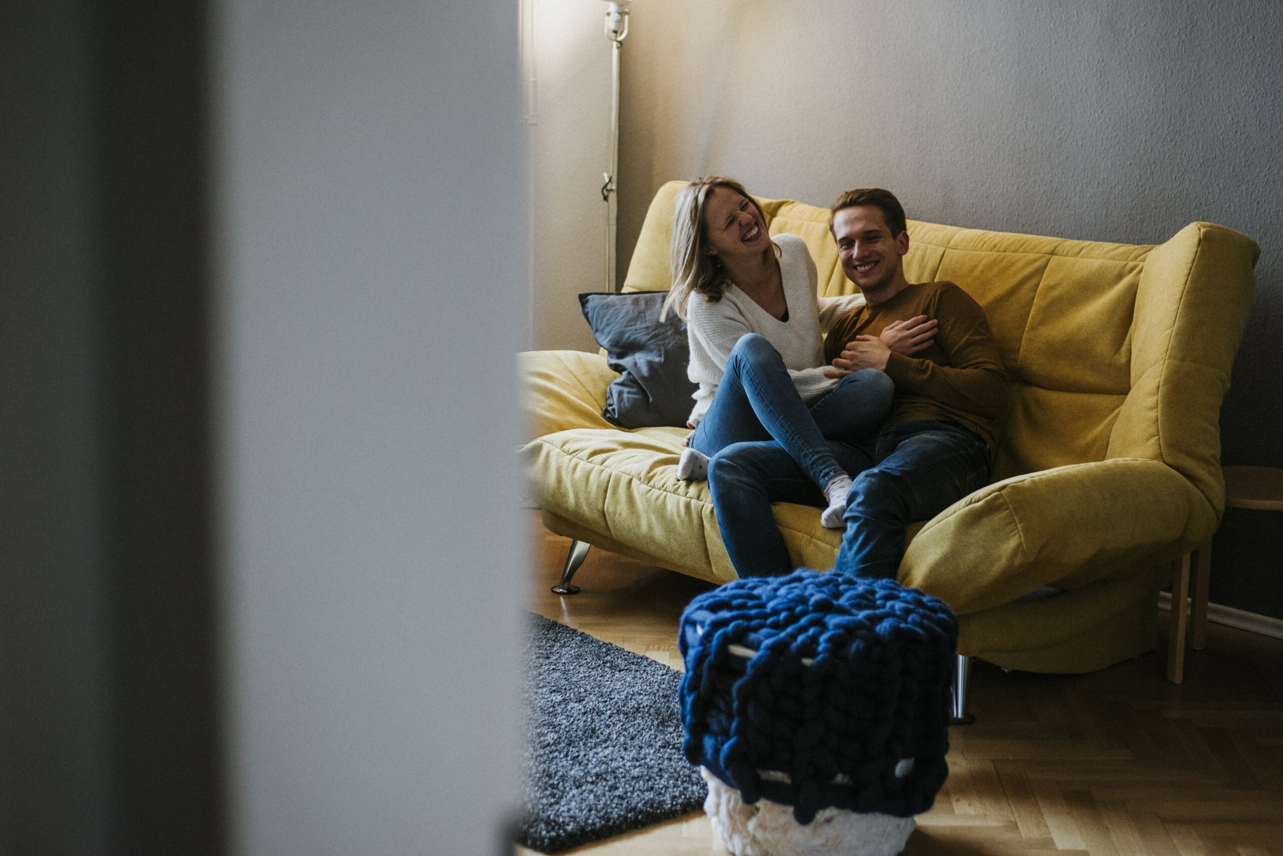 Paar lachend auf gelbem Sofa, blauer Strickhocker im Vordergrund, gemütlich eingerichteter Raum.