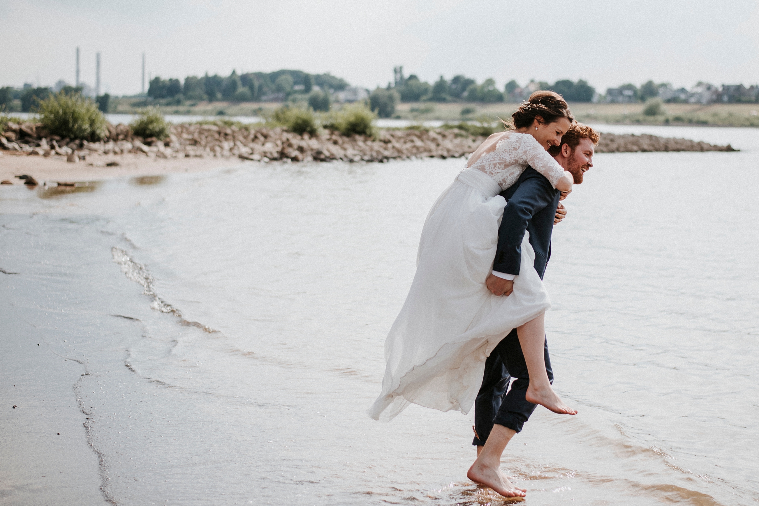 Mann trägt Frau in weißem Kleid auf dem Rücken am Wasser entlang, Bäume und Gebäude im Hintergrund.