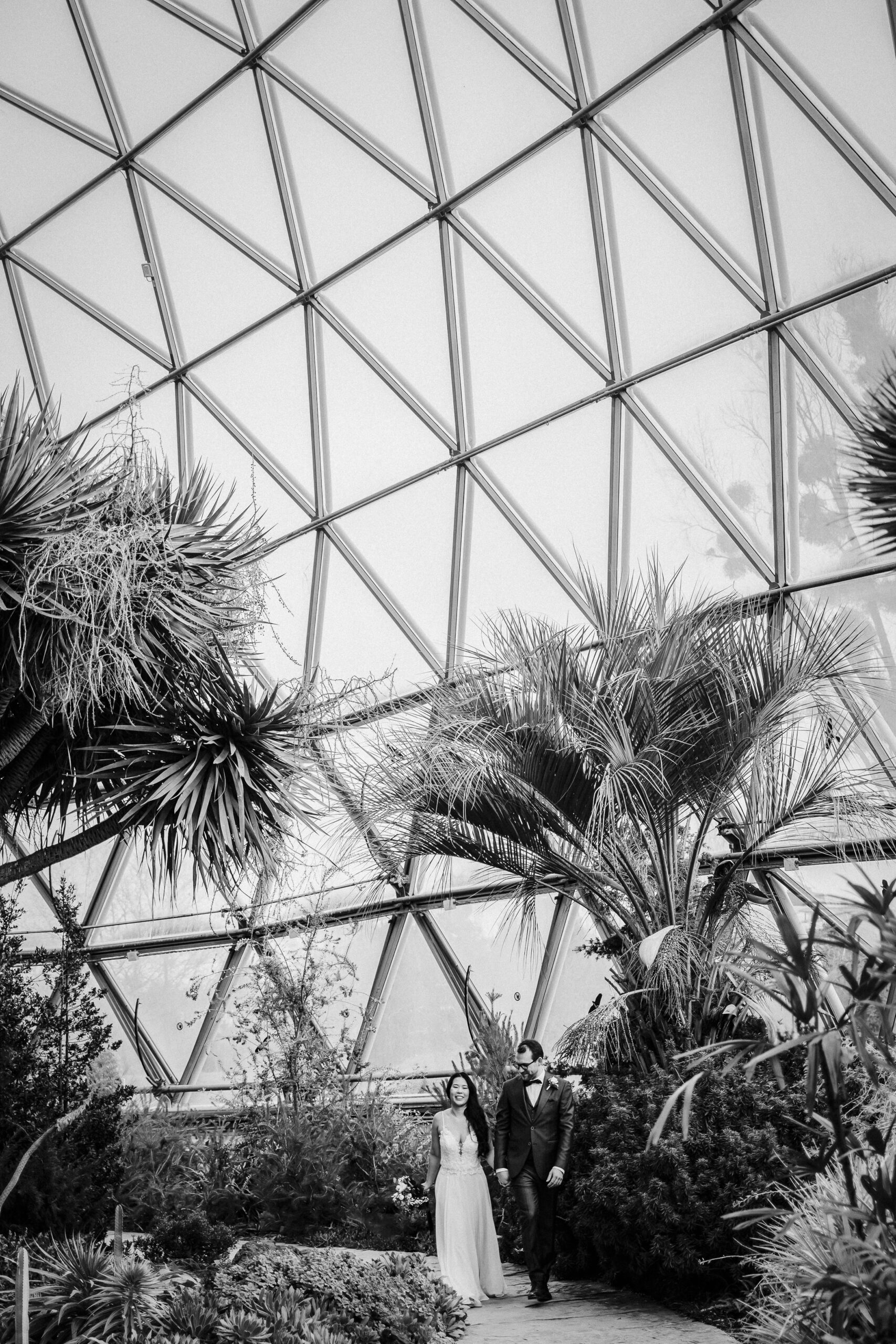 Brautpaar im Gewächshaus mit üppiger Vegetation und geometrischem Glasdach, Schwarzweiß-Fotografie.