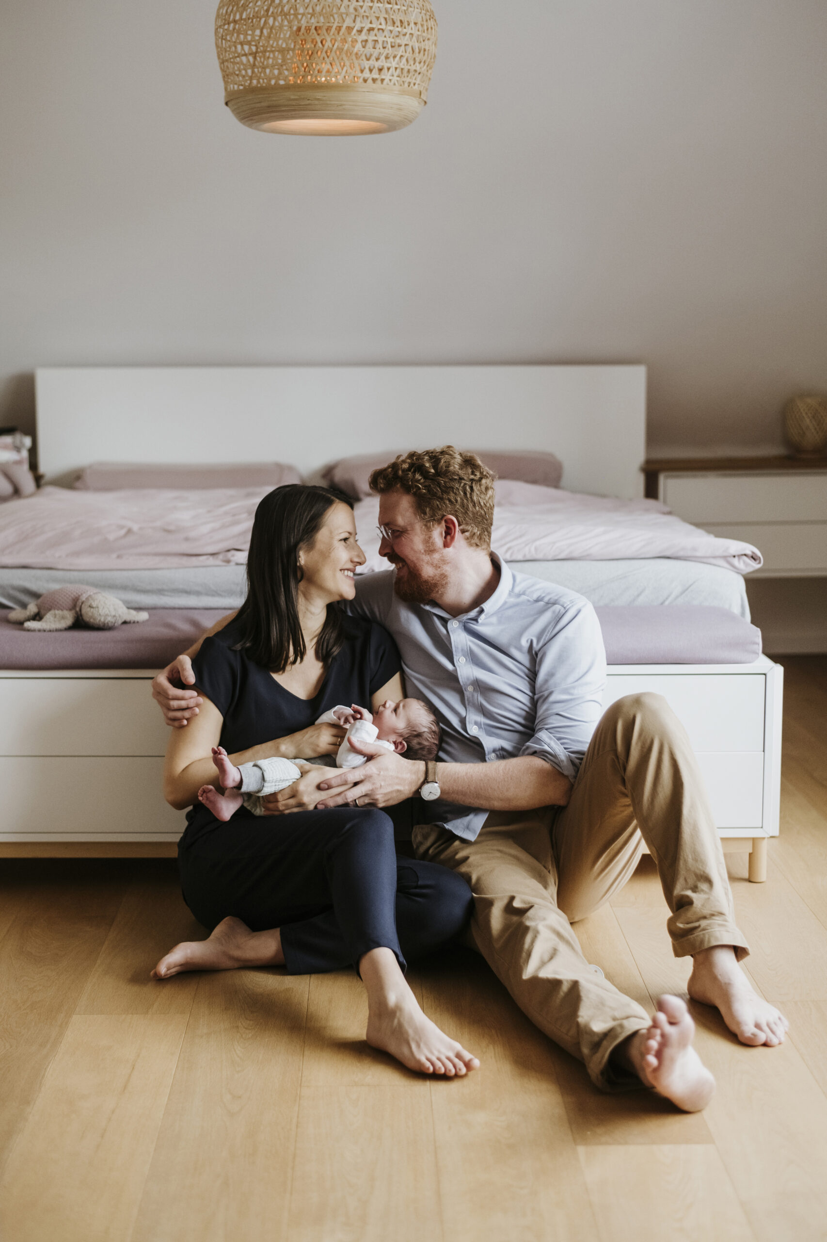 Paar auf Holzboden vor einem Bett hält lächelnd ein schlafendes Baby; gemütliche Umgebung.