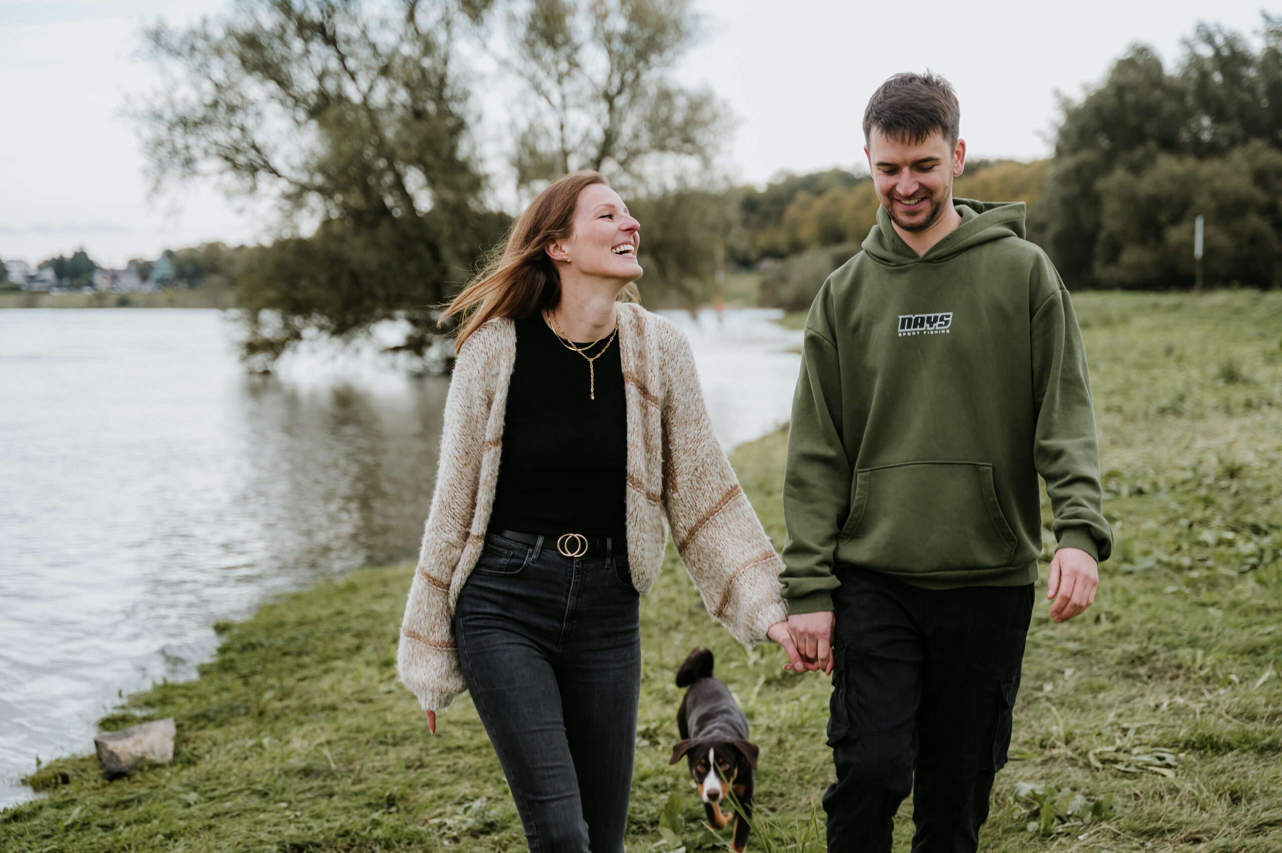 Paar spaziert mit Hund am Flussufer entlang, umgeben von Bäumen und Wasser im Hintergrund.