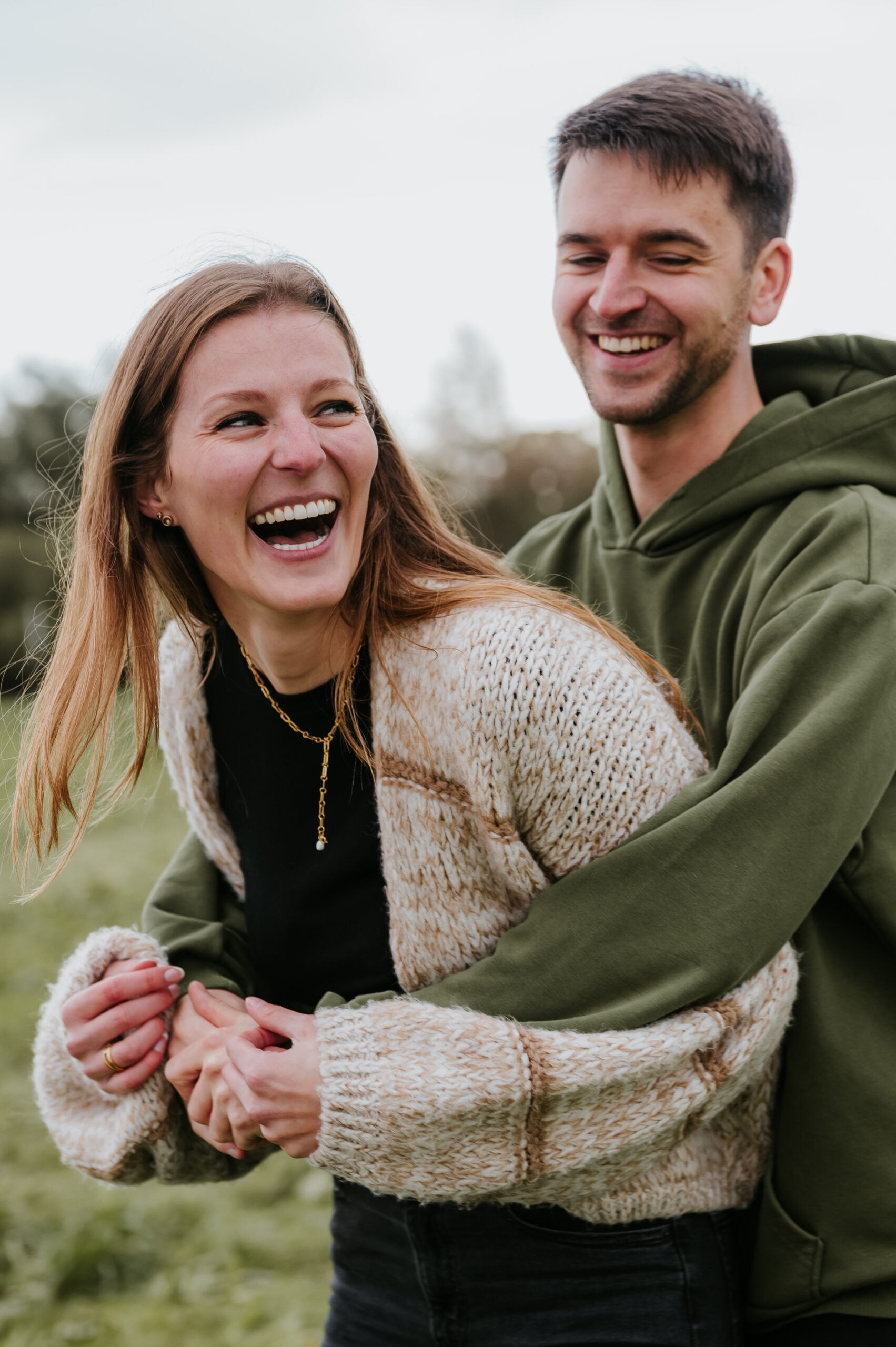 Paar lächelt entspannt im Freien; Frau in Strickpullover, Mann in olivgrünem Hoodie.