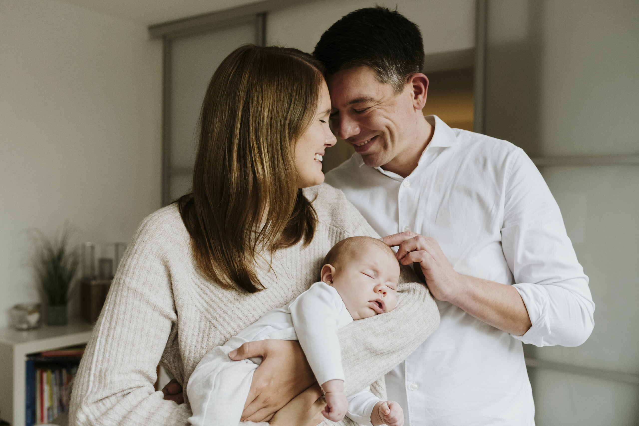 Paar hält schlafendes Baby in gemütlichem Raum, beide Eltern lächeln einander an.