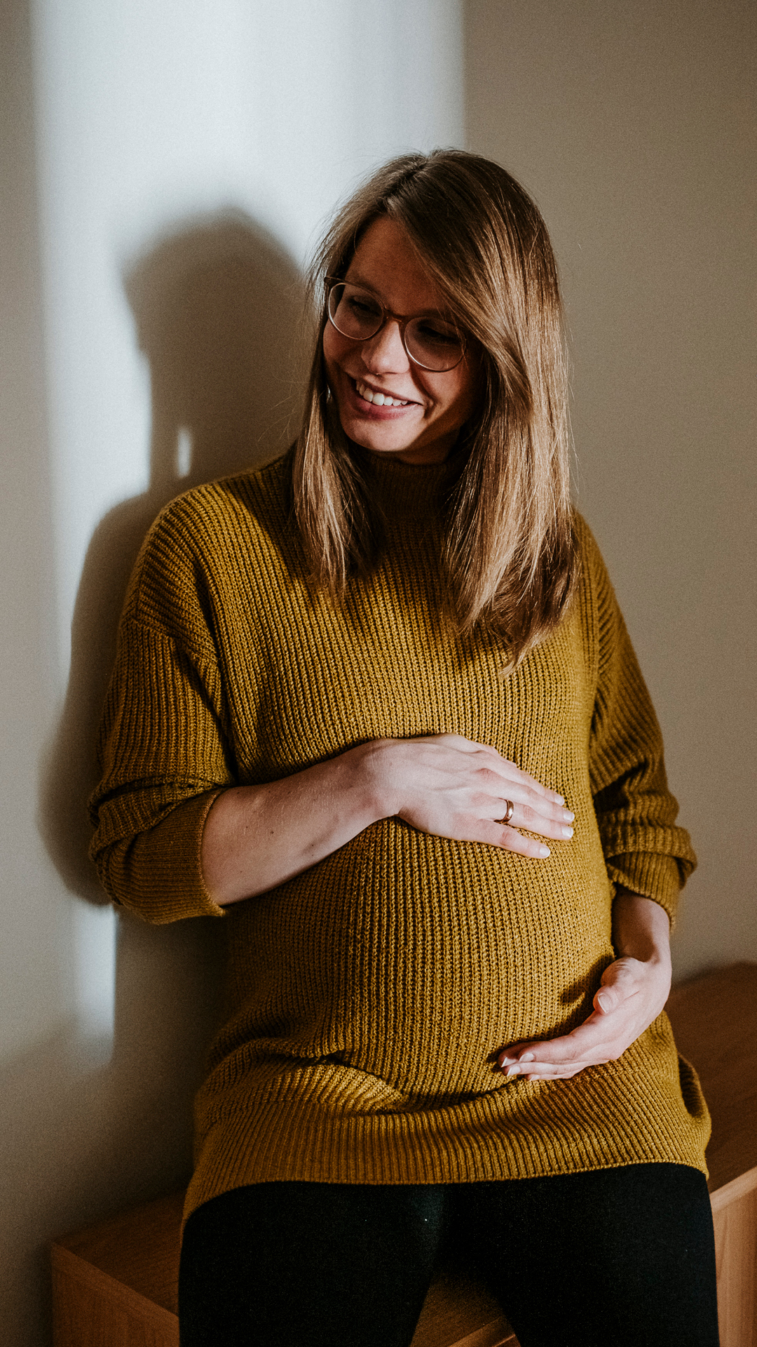 Schwangere Frau im senfgelben Pullover, hält lächelnd ihren Bauch und trägt eine Brille.