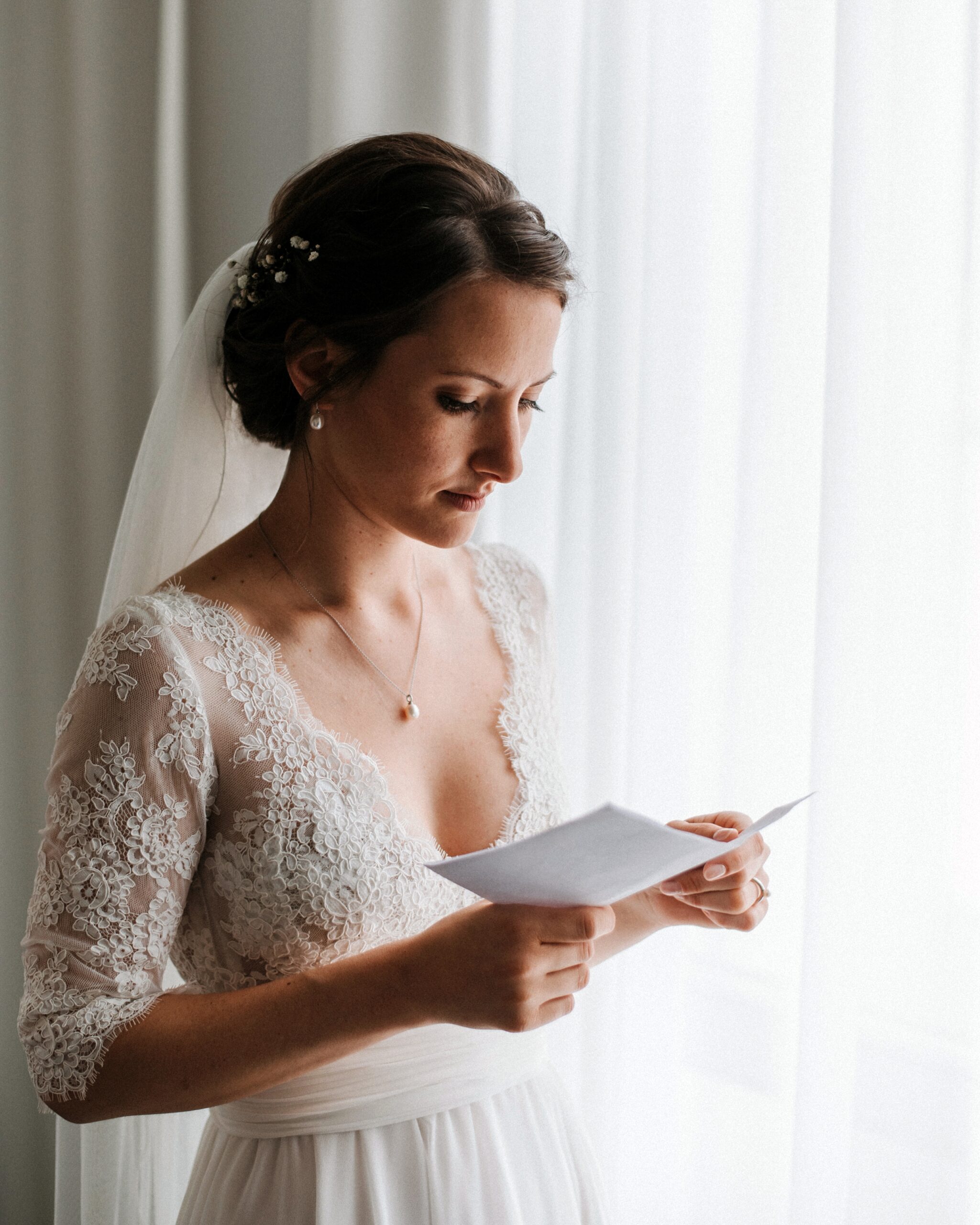 Frau im Spitzenhochzeitskleid liest ihr Eheversprechen am Fenster, trägt einen Schleier und Haarschmuck.
