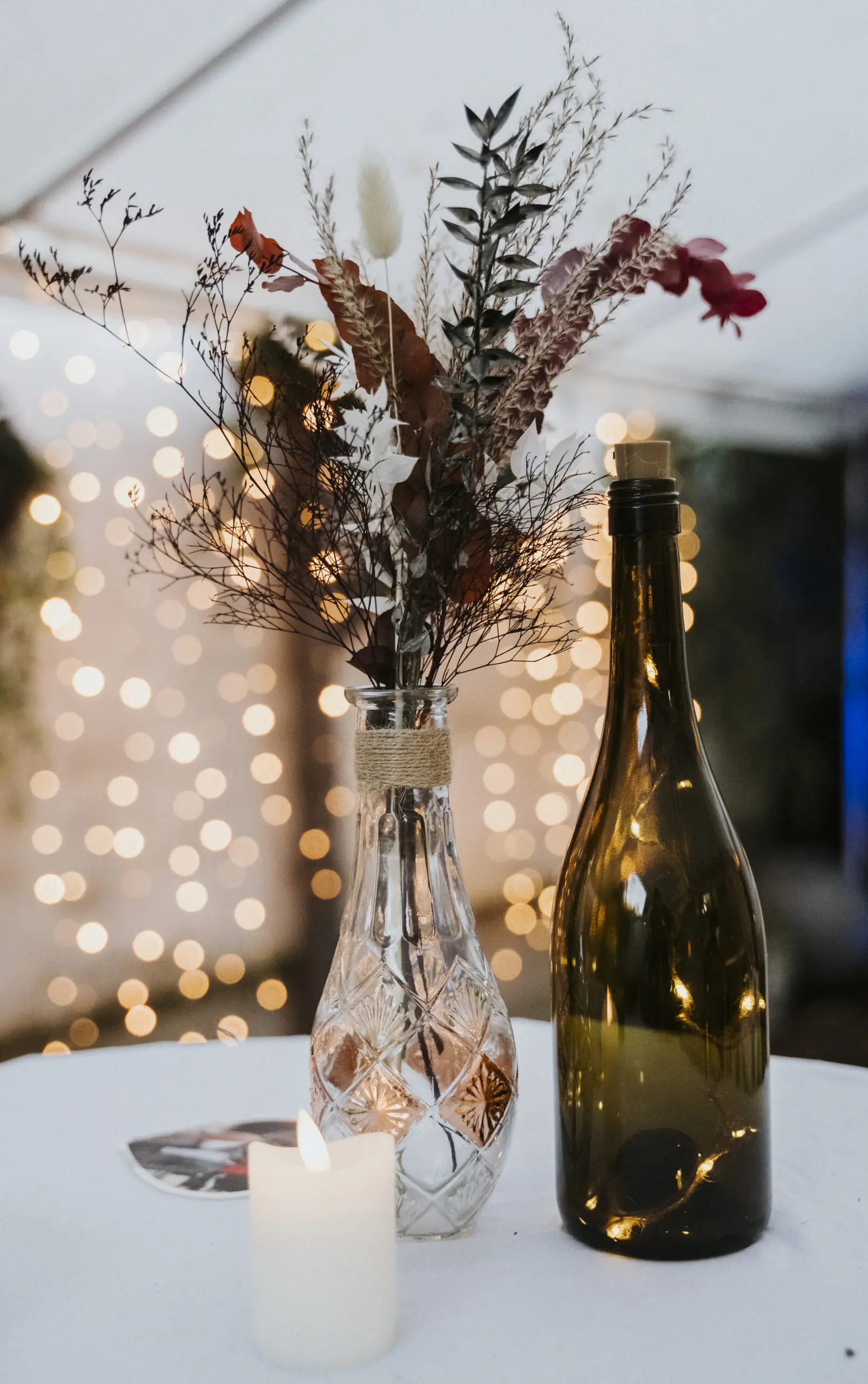 Dekorativer Tisch mit Kristallvase, Trockenblumen, leere Flasche, Kerze, verschwommene Lichter.