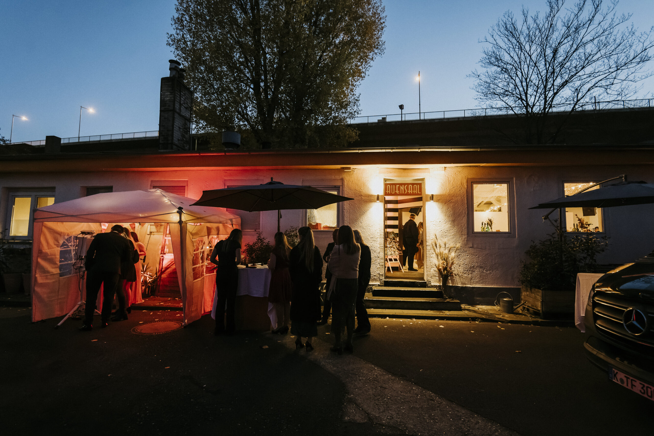 Abendveranstaltung vor Gebäude, beleuchtetes Zelt und "Avesaal"-Eingang, versammelte Menschen.