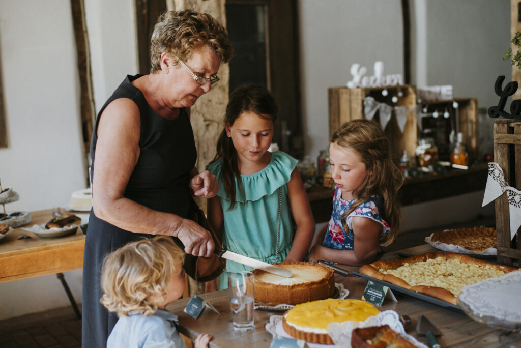Ältere Frau schneidet Kuchen, drei Kinder schauen zu; Tisch mit verschiedenen Backwaren.