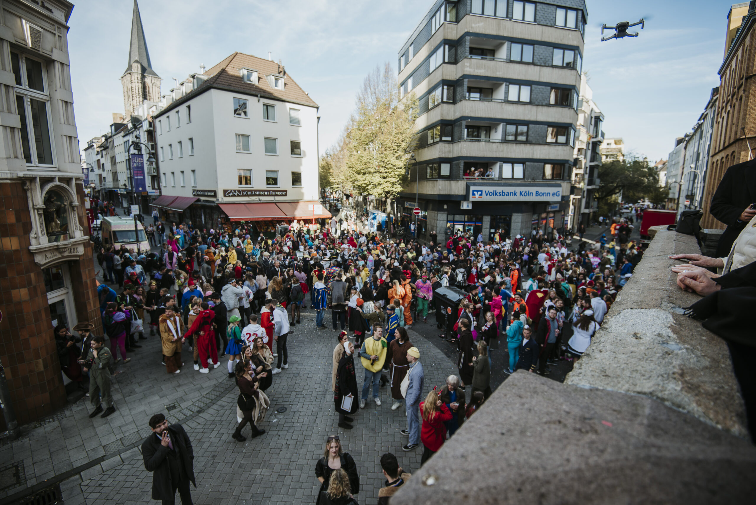 Bunte Menschenmenge in Kostümen auf städtischem Platz, Gebäude und Drohne im Hintergrund.