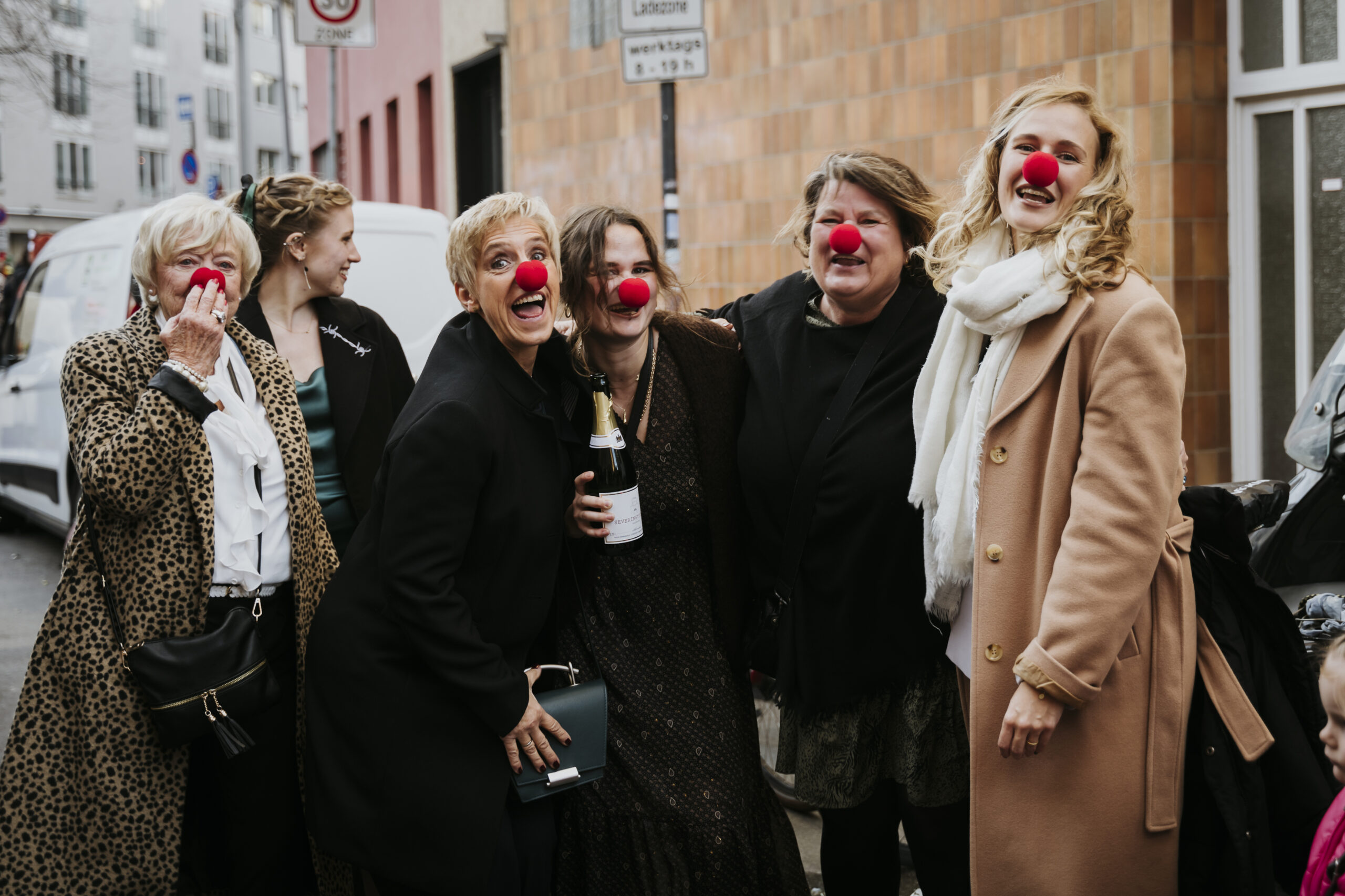 Sechs Personen mit roten Clownsnasen lächeln auf der Straße, eine hält eine Flasche.