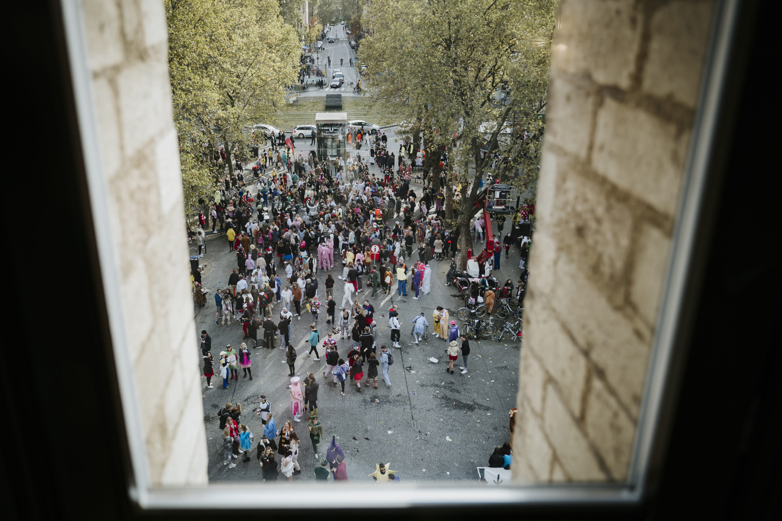 Menschenmenge in bunten Kostümen auf städtischem Platz, von einem Fenster aus gesehen.