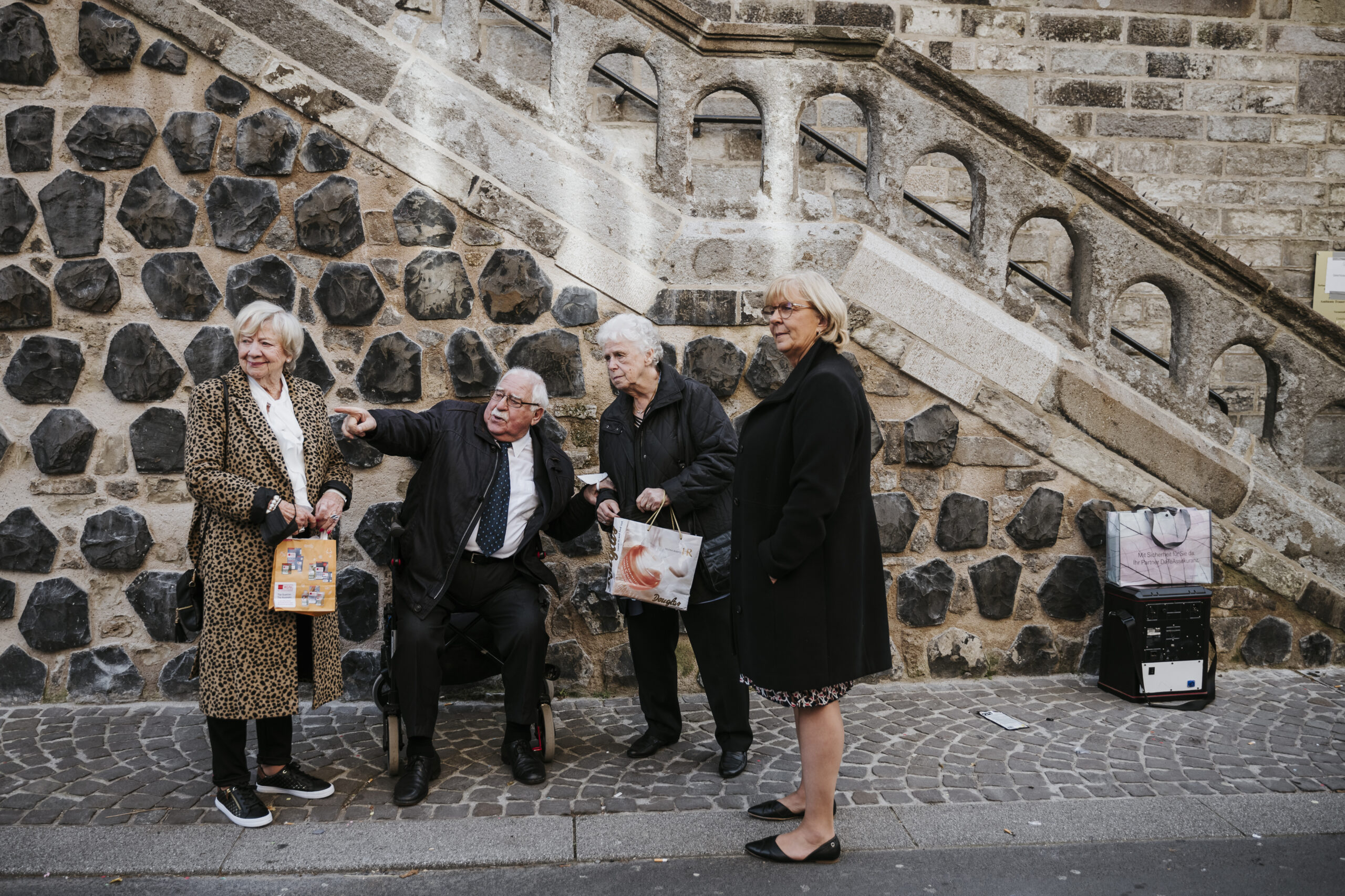 Ältere Menschen vor Steinmauer auf gepflastertem Weg, eine Person im Rollstuhl, zeigt gestikulierend.