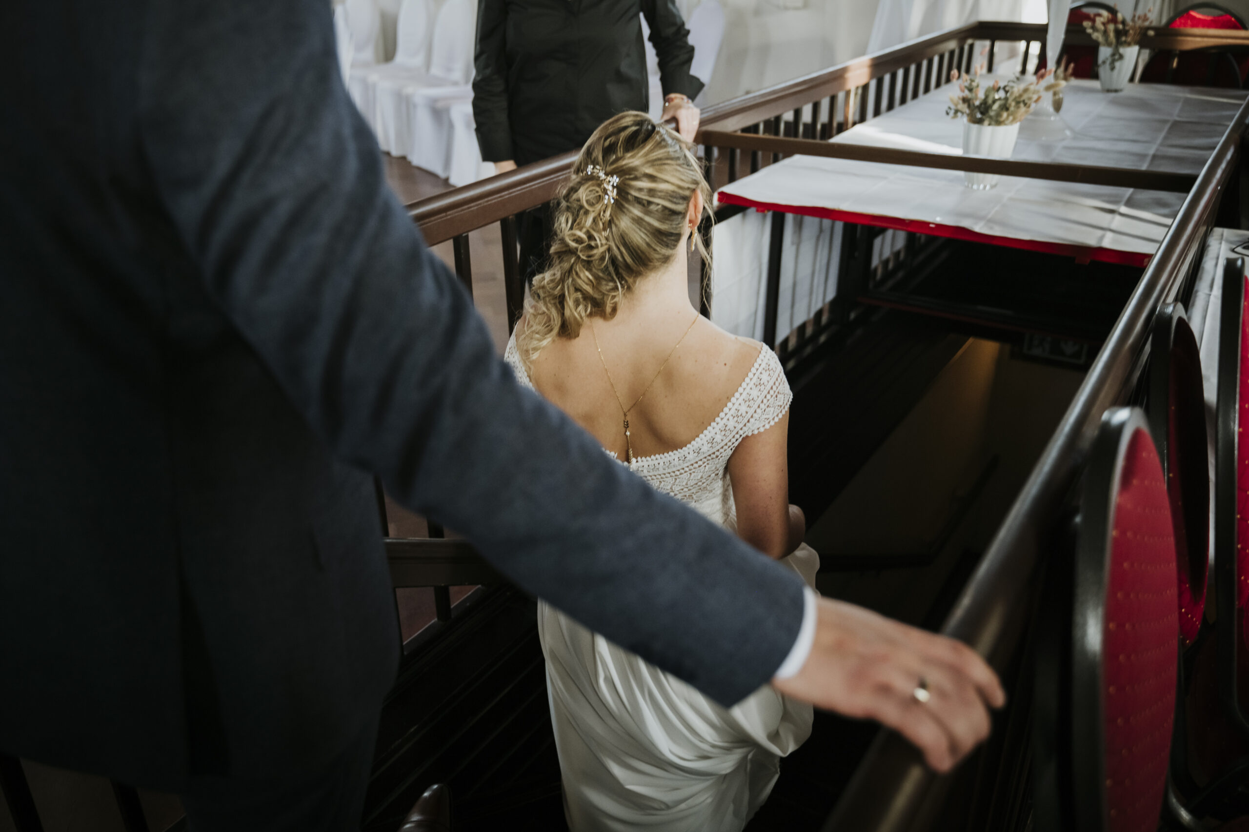 Elegant bridal gown-clad woman descends stairs, accompanied by suited person, white chairs nearby.