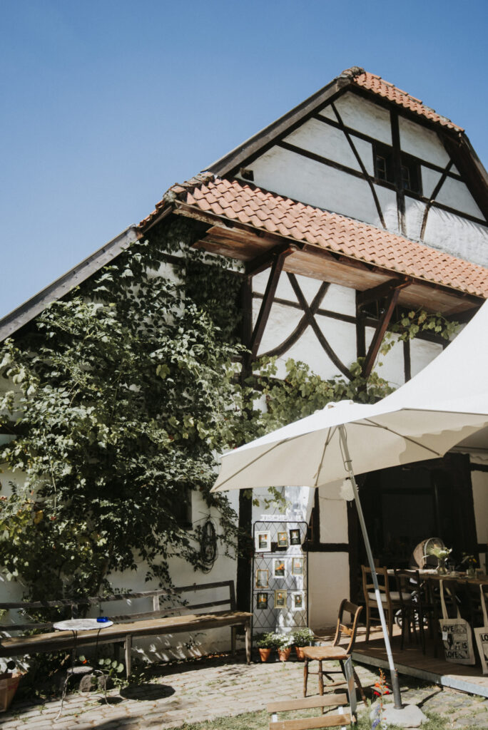 Fachwerkhaus mit bewachsenem Giebel, Sonnenschirm, Tischen, Stühlen, Pflanzen und Dekorationen.