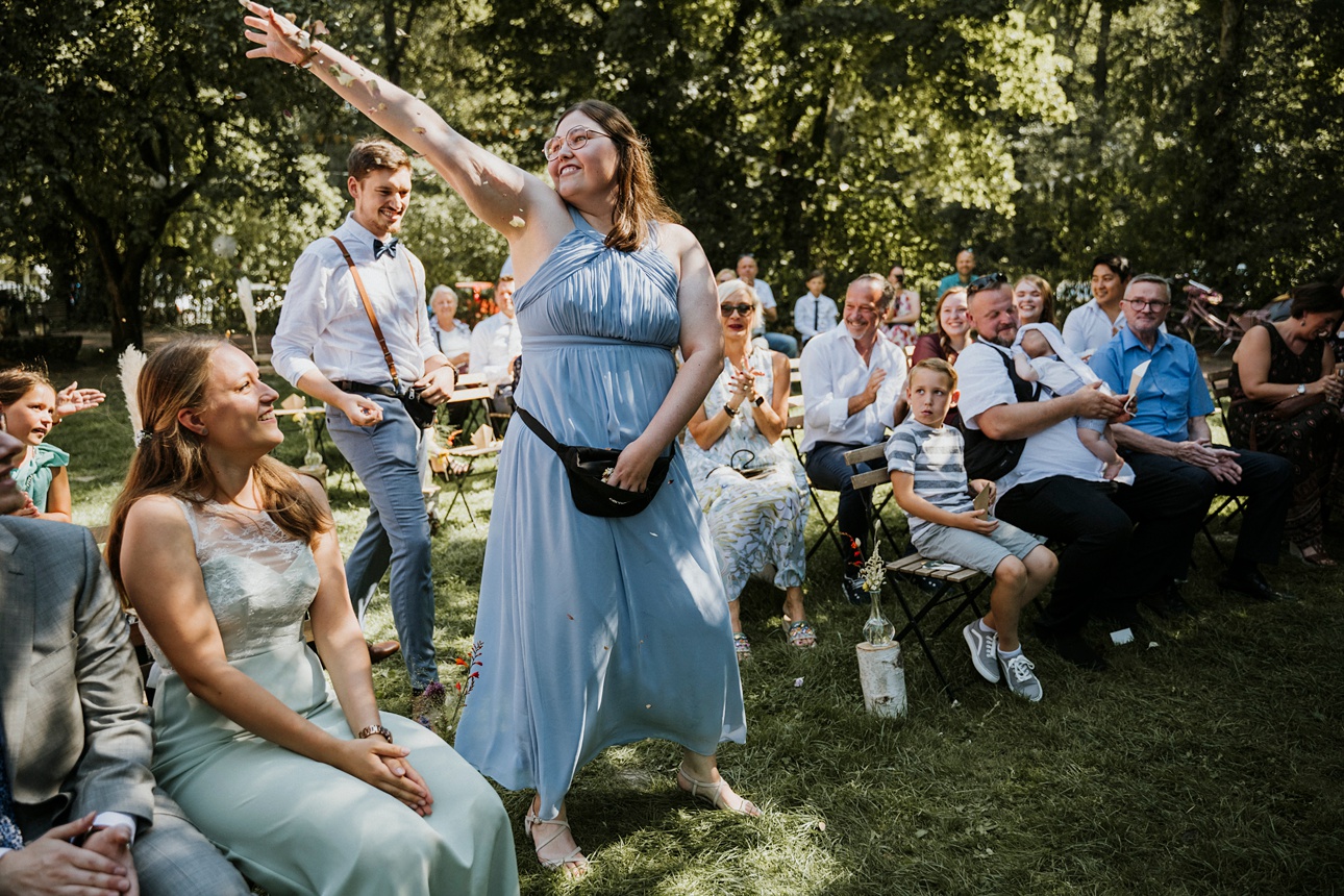 Frau im blauen Kleid wirft bei Outdoor-Event etwas in die Luft, umgeben von sitzenden Gästen.