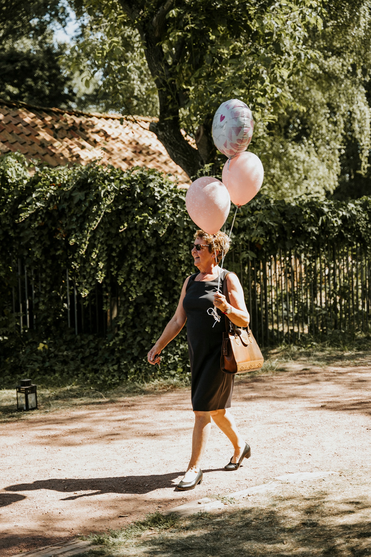 Frau spaziert auf sonnigem Weg mit drei rosa Ballons, Sonnenbrille und Ziegeldach Gebäude.