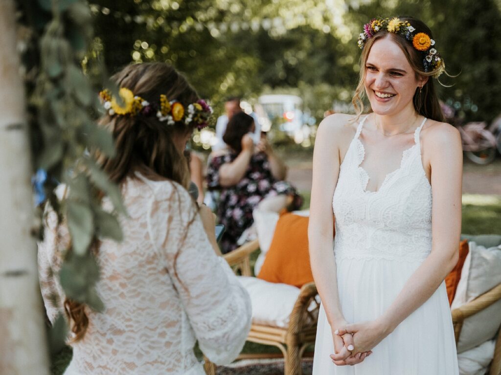 Frau im weißen Kleid und Blumenkranz lächelt im Garten; unscharfe Person im Vordergrund.