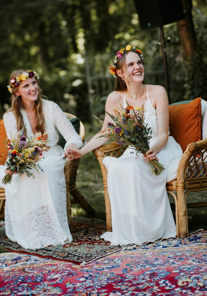 Frauen in weißen Kleidern mit Blumenkränzen und Sträußen, auf Rattan-Stühlen im Garten.