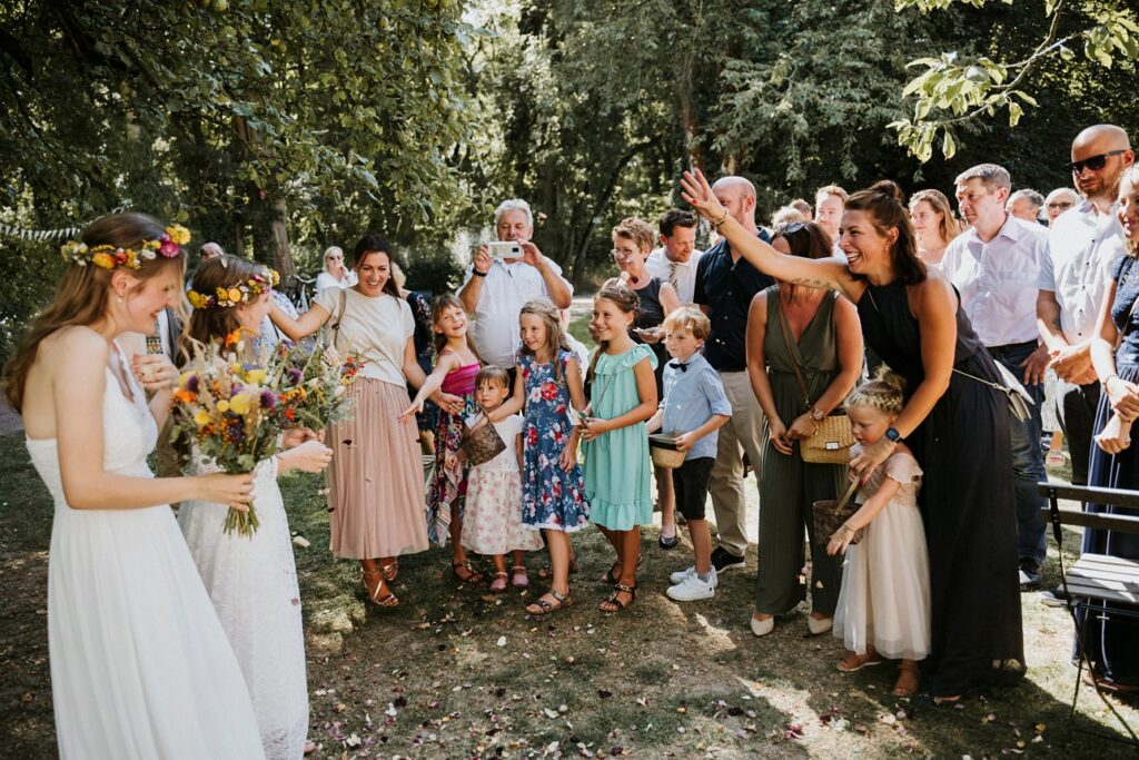 Fröhliche Hochzeitsgesellschaft im Freien mit Blumen, Kindern und grüner Vegetation.