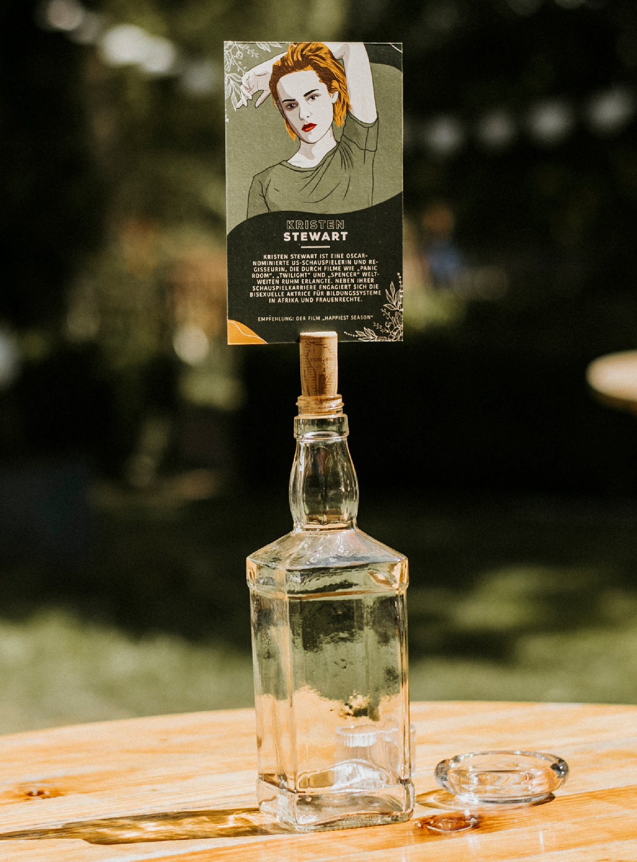 Leere Glasflasche mit Karte von Person auf Holztisch, unscharfer grüner Hintergrund.