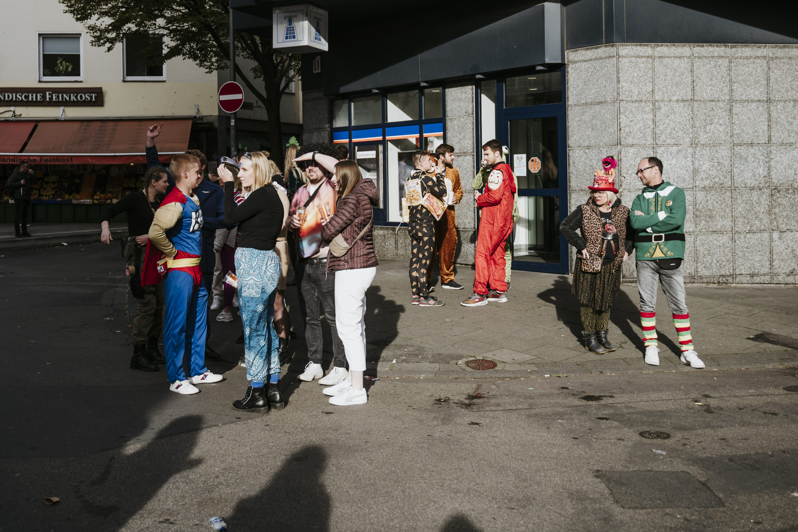 Gruppe in bunten Kostümen auf sonniger Straße, einige unterhalten sich, Geschäft im Hintergrund.