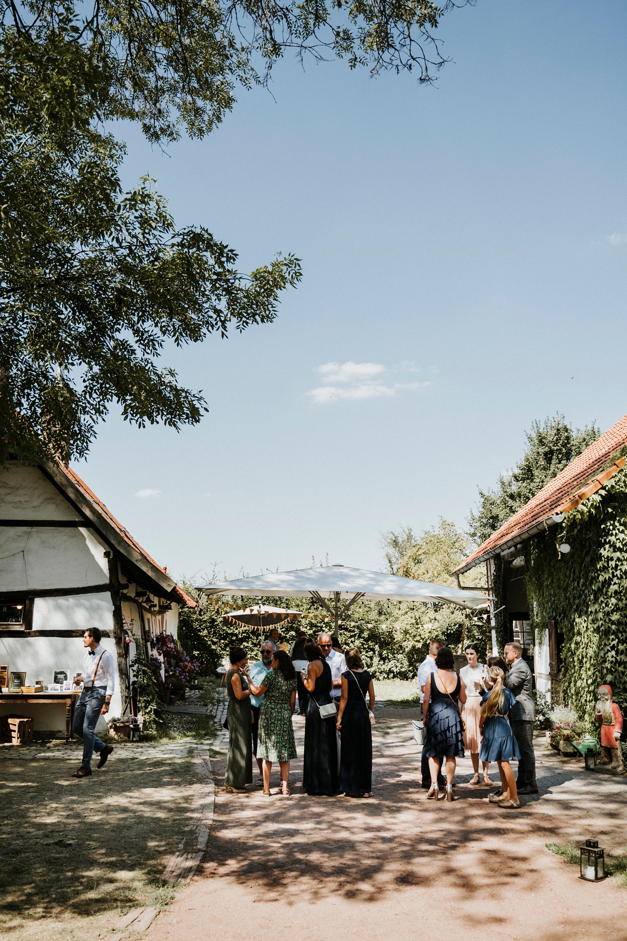 Gruppe im Freien unter Sonnenschirm, umgeben von Efeu-bedeckten Gebäuden, Bäume im Hintergrund.