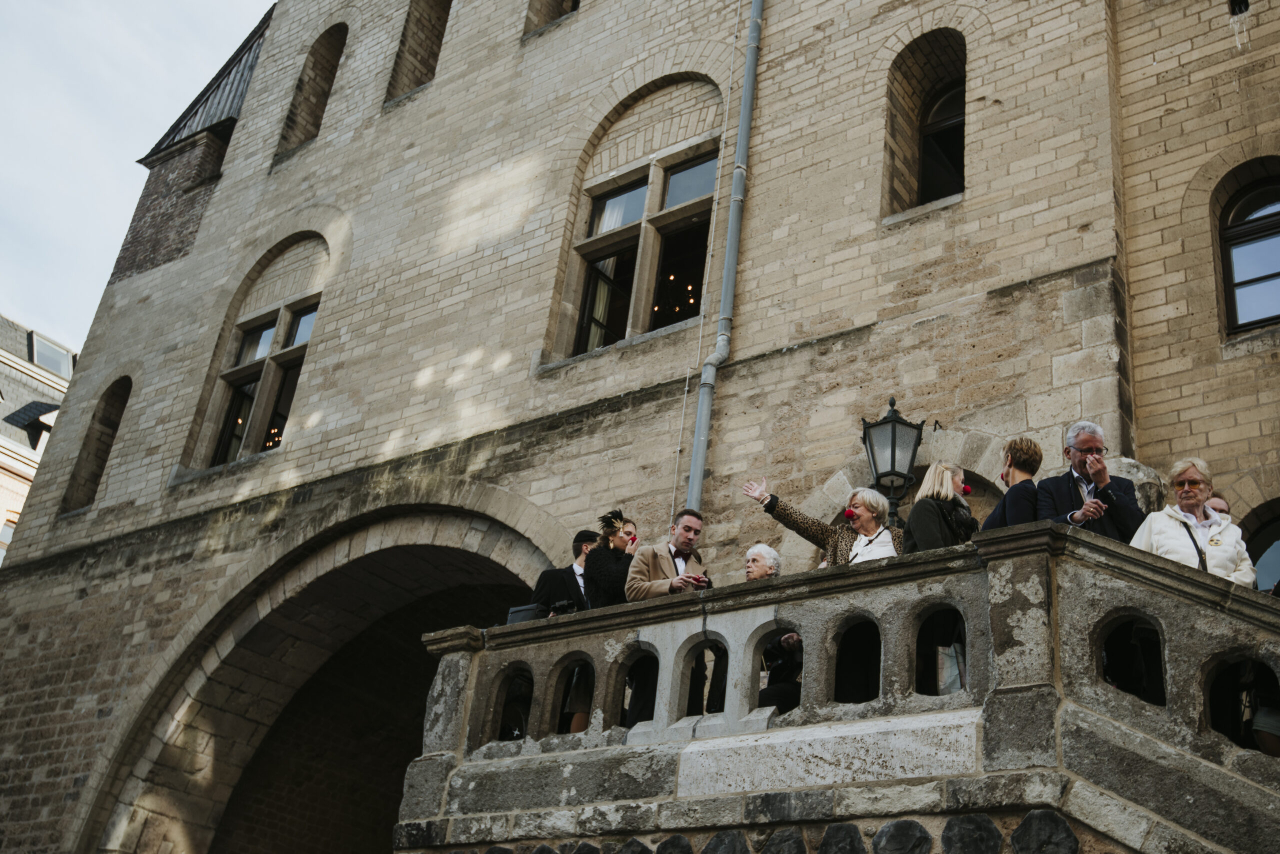 Gruppe auf historischem Balkon, einige mit roten Clownsnasen, steinernes Gebäude im Hintergrund.