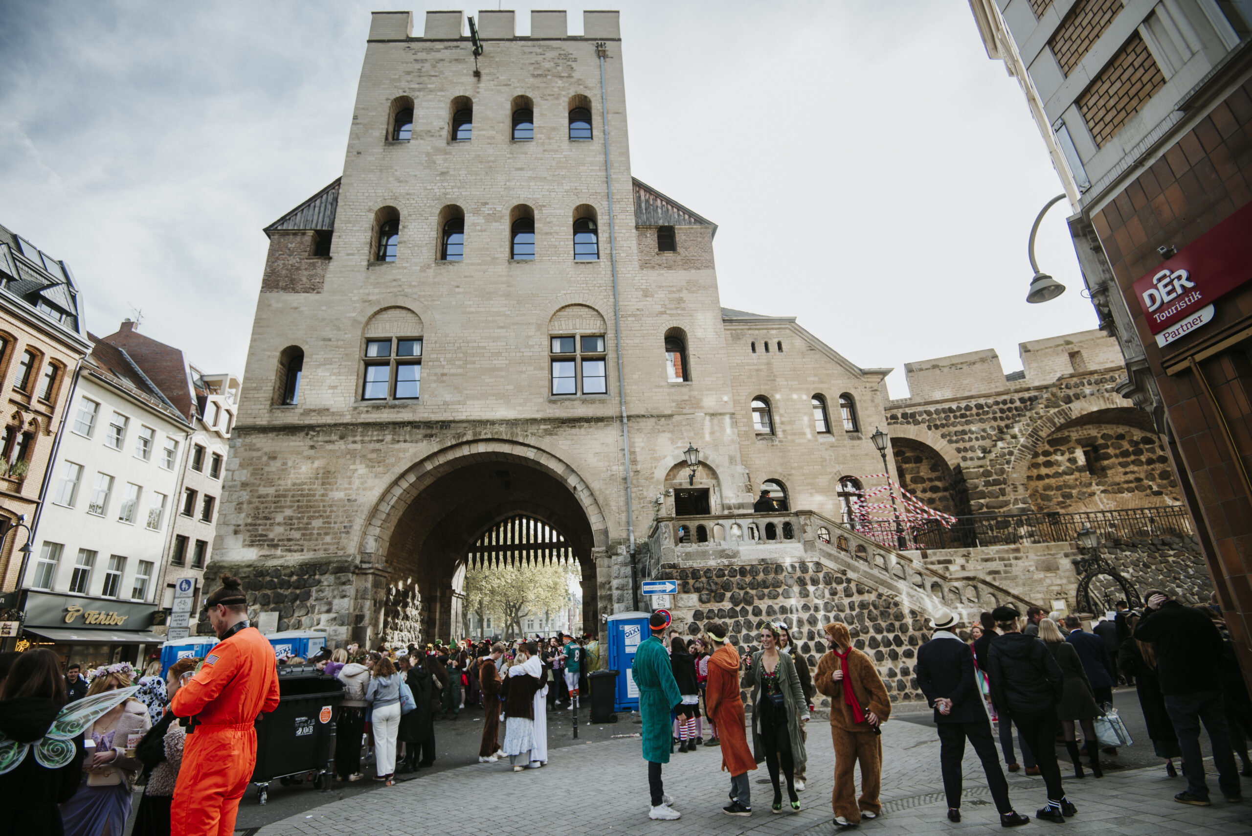 Historisches Stadttor mit versammelter Menschenmenge in bunten Kostümen, Gebäude im Hintergrund.