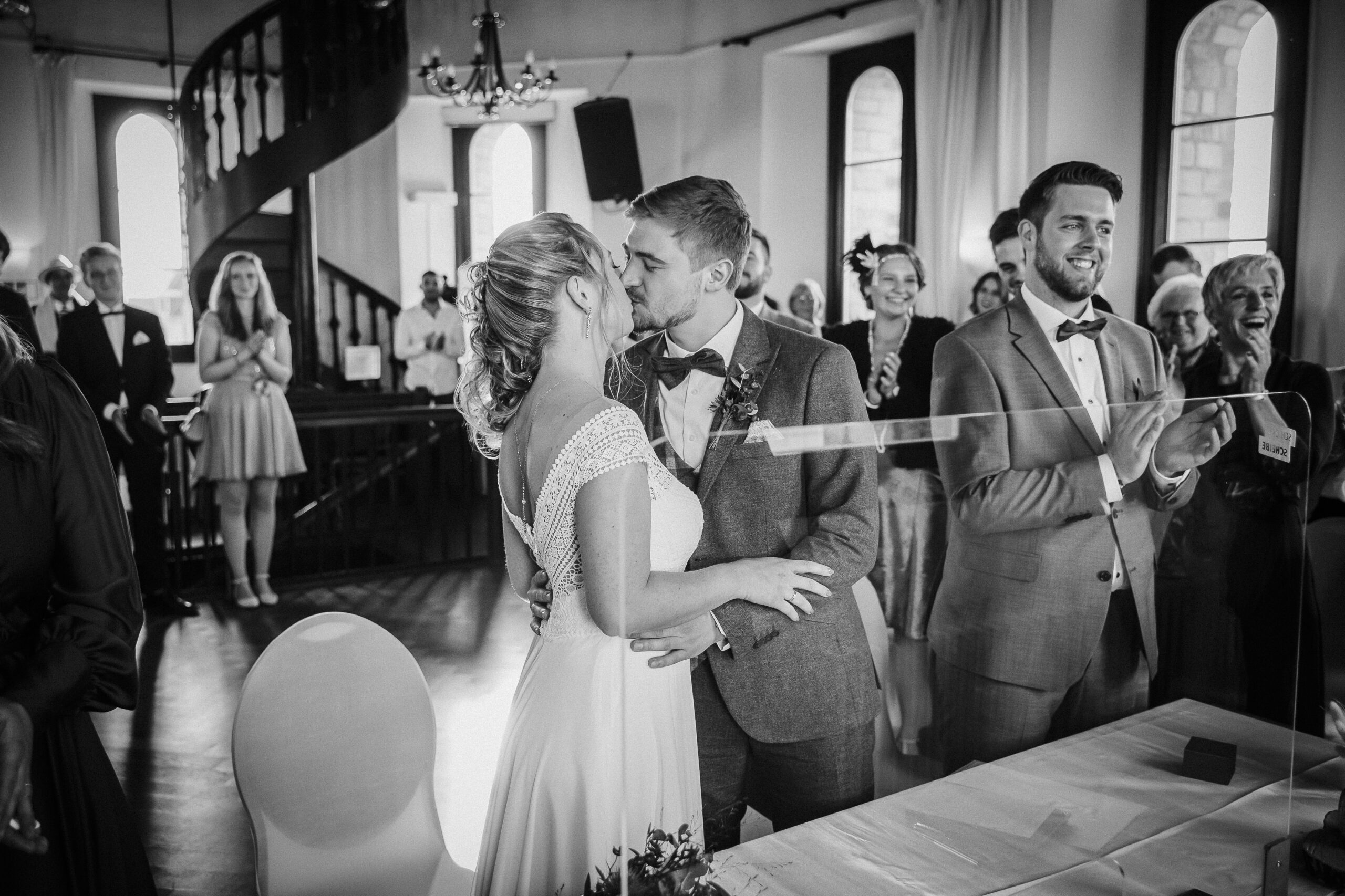 Hochzeitspaar küsst sich vor applaudierenden Gästen in festlichem Raum mit Wendeltreppe.