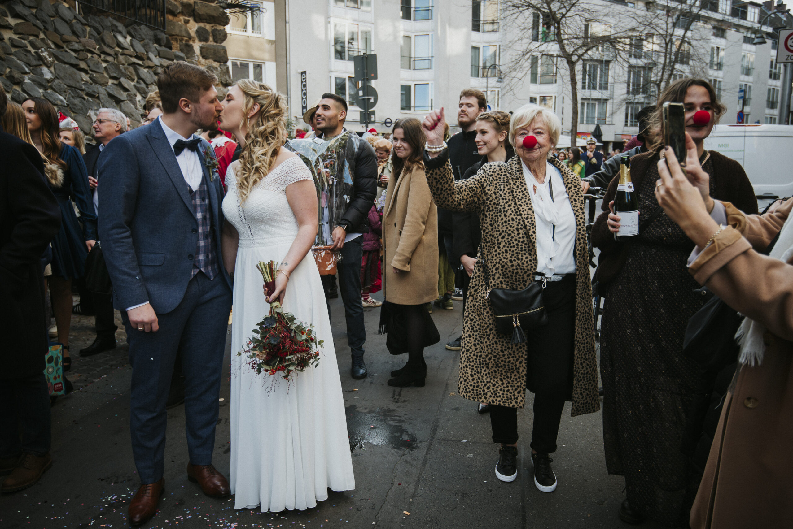 Hochzeitspaar auf Straße mit Clownsnasen, lächelnden Gästen und Gebäuden im Hintergrund.
