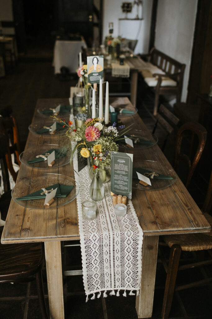 Holztisch mit gehäkeltem Tischläufer, Glasgeschirr, grünen Servietten, Blumen und Kerzen.