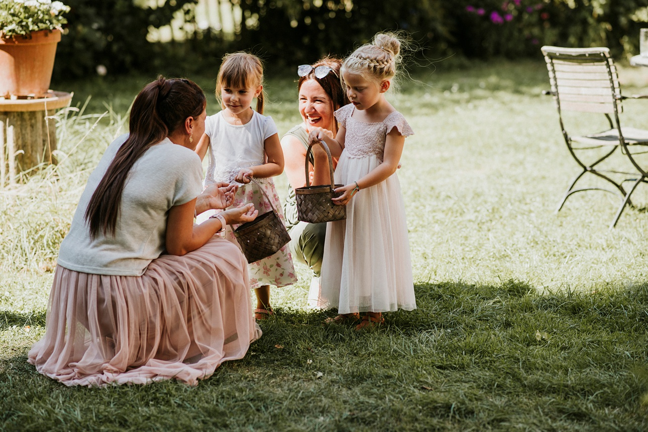 Kinder in festlicher Kleidung mit Körben auf Wiese, zwei Frauen interagieren mit ihnen.