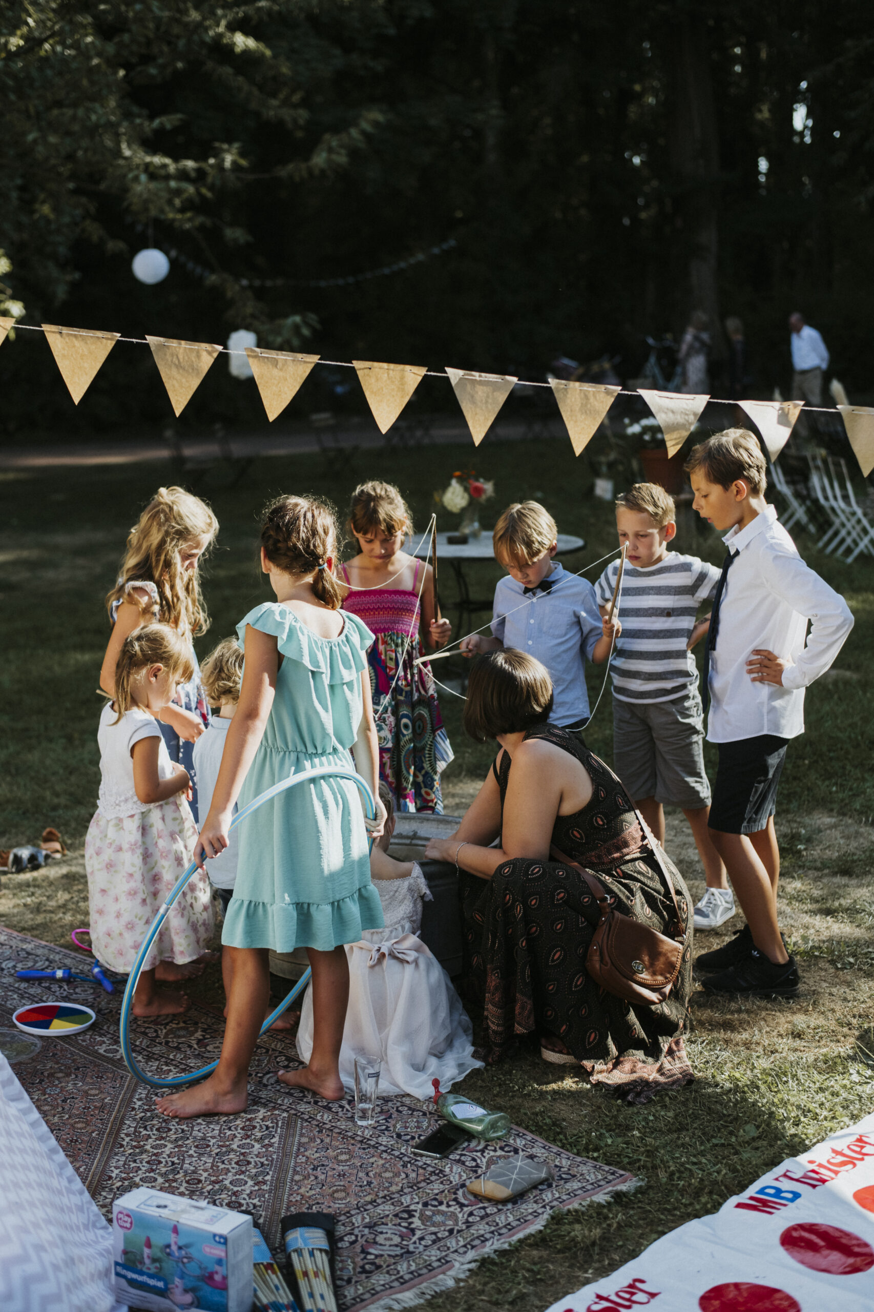 Kinder spielen im Freien mit einem Erwachsenen auf einem Rasen mit Teppich und Wimpelkette.