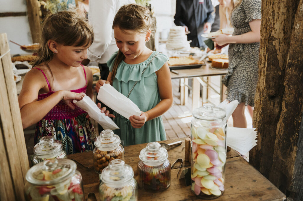 Zwei Kinder füllen Süßigkeiten in Tüten an einem Tisch mit Gläsern; Menschen am Buffet im Hintergrund.