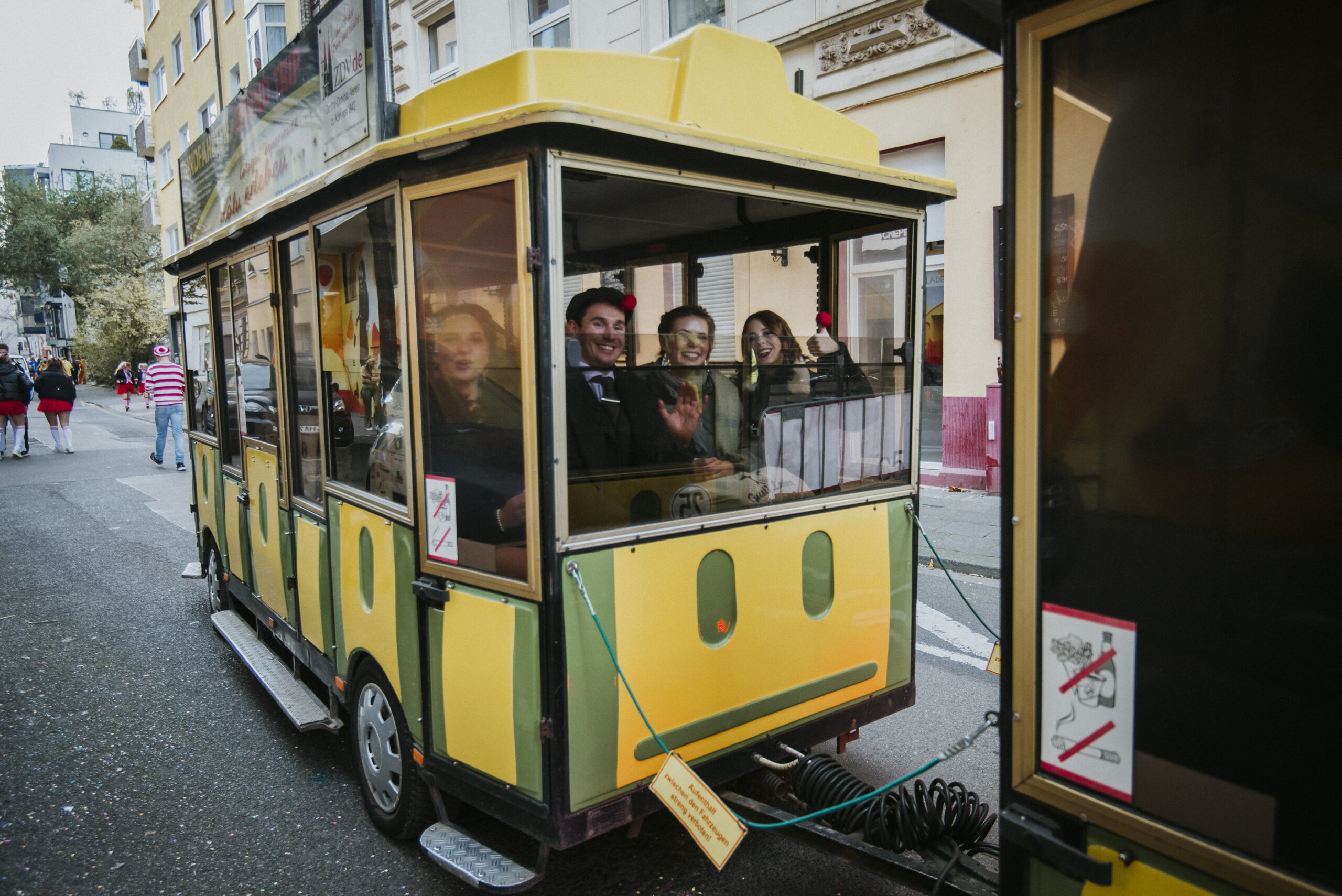 Kleine gelbe Straßenbahn mit fröhlichen Fahrgästen in einer Stadt, Rauch- und Trinkverbot.
