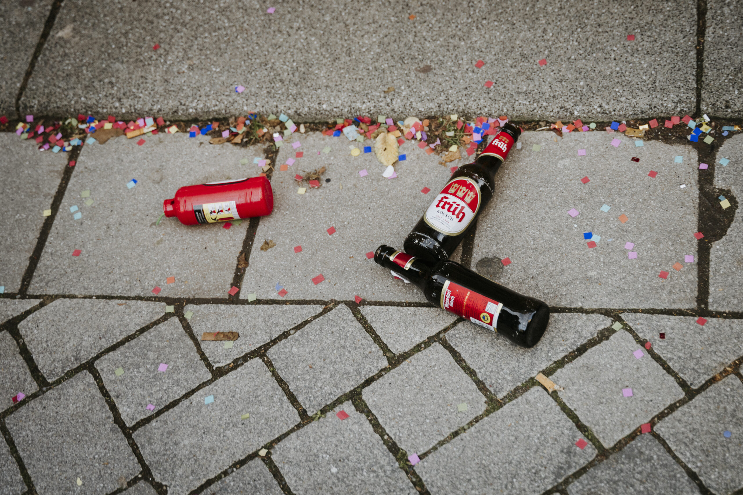 Leere Glasflaschen und Plastikebgeflasche auf Gehweg mit buntem Konfetti verstreut.