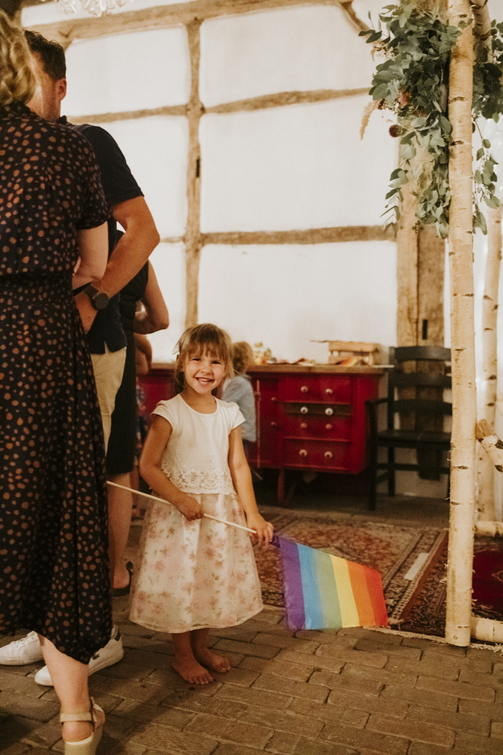 Mädchen im Kleid mit Regenbogenflagge in Raum mit Holzbalken, roter Kommode und Teppich.