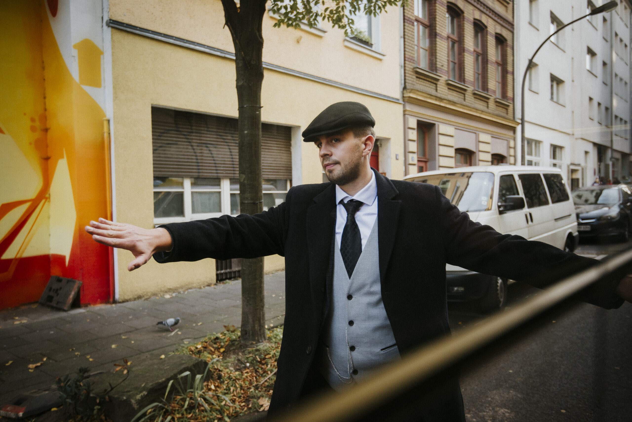 Mann in Anzug hebt Arm auf Straße, bemalte Wand, geparkte Autos, Wohngebäude im Hintergrund.