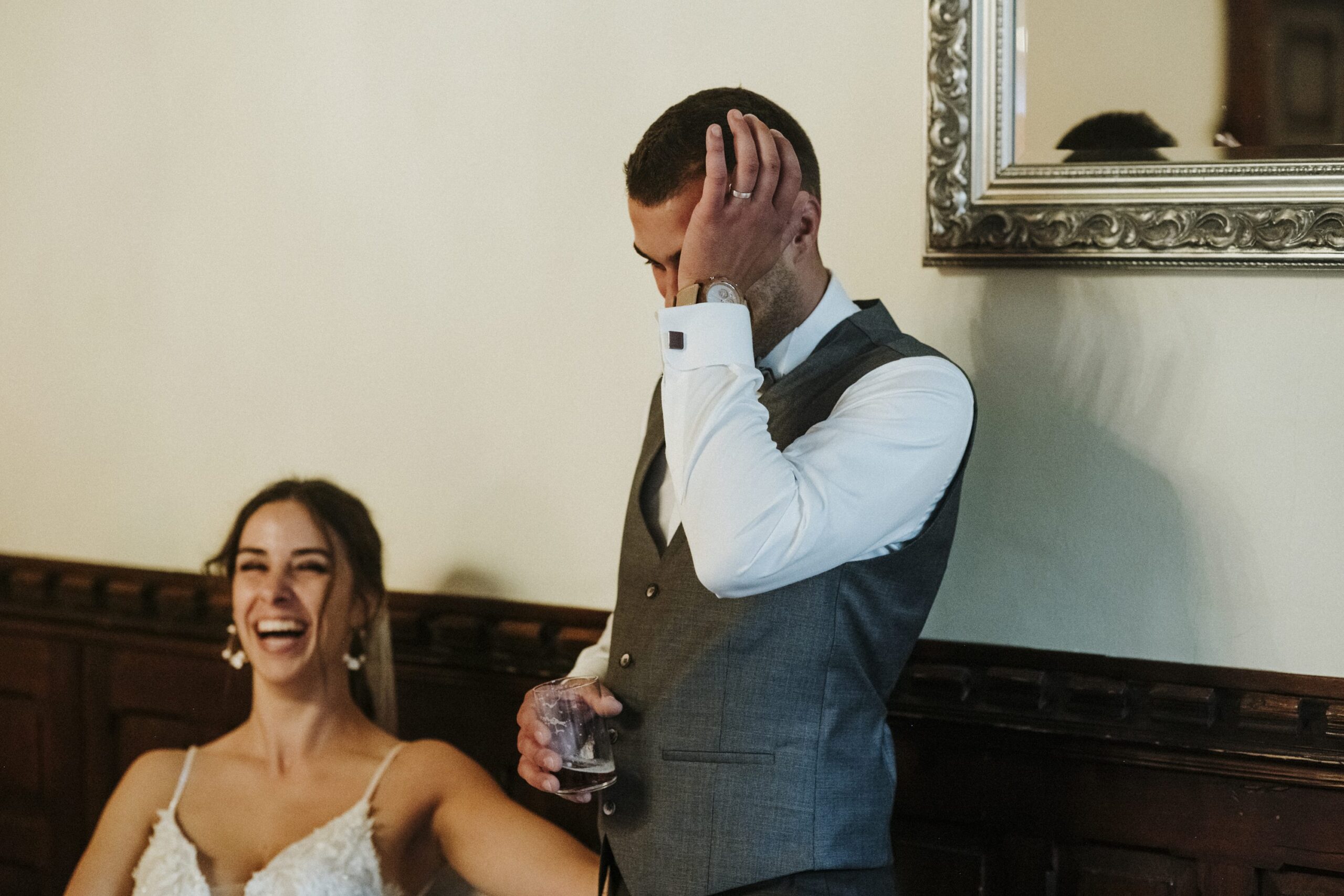 Mann in Anzug verdeckt Gesicht mit Hand, Frau im Hochzeitskleid lacht, Spiegel im Hintergrund.