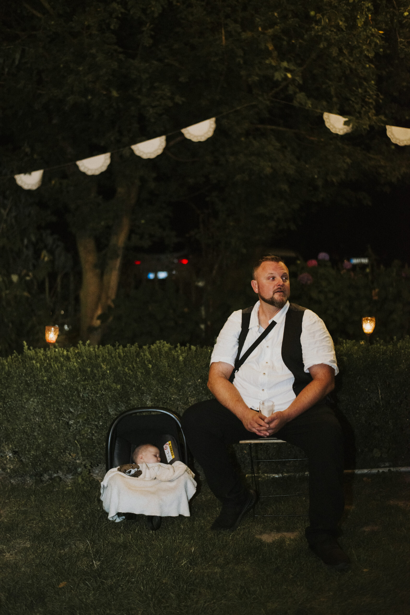Mann im Freien auf Stuhl neben schlafendem Baby, Bäume und Lichterkette im Hintergrund.