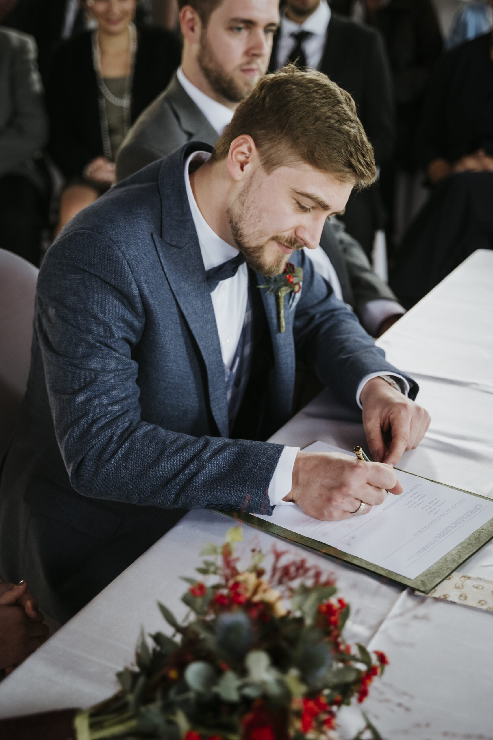 Mann unterschreibt Dokument im Anzug am Tisch mit Blumen, Hintergrund mit sitzenden Personen.